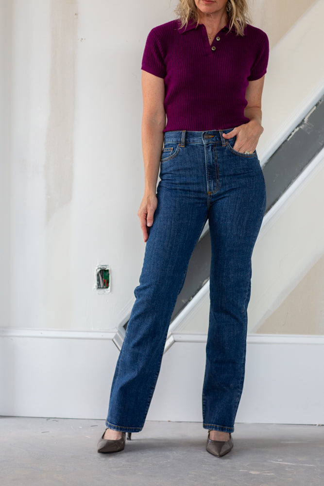 Person wearing a purple shirt and blue jeans standing against a white wall.