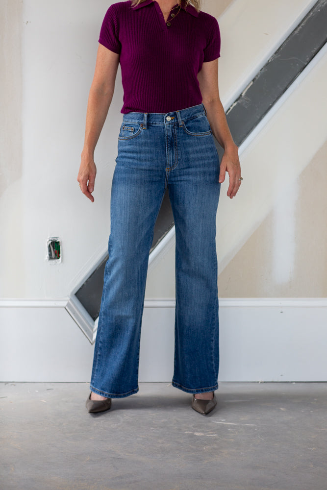Person wearing a maroon top and blue jeans standing against a white wall.