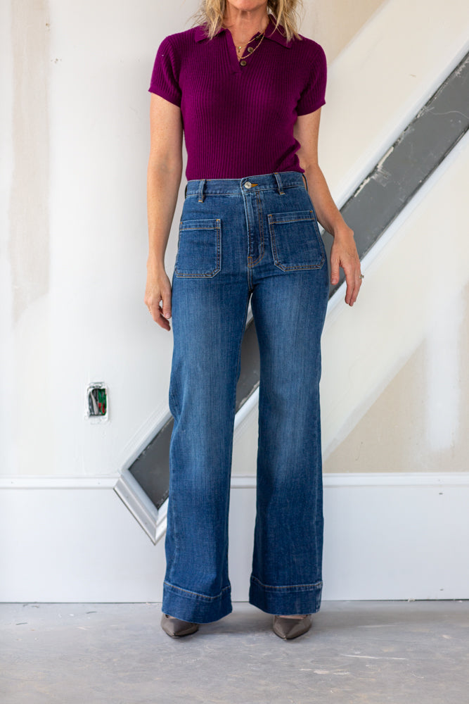 Person wearing a purple shirt and blue jeans standing against a white wall.