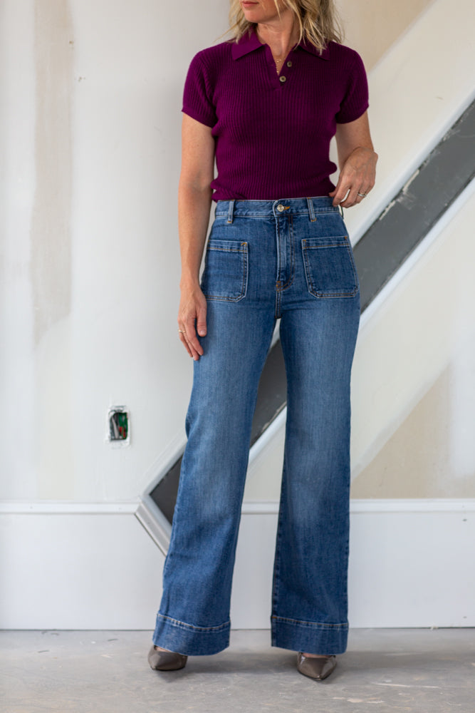 Person wearing a purple polo shirt and blue jeans standing against a white wall.