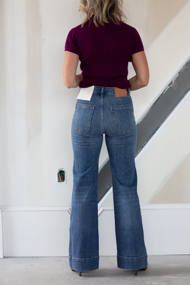 Person wearing blue jeans and a maroon top standing in a minimal indoor setting.