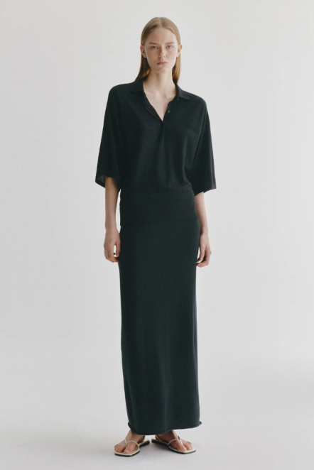 A model stands against a light gray studio background wearing a long black collared dress with short sleeves and flat sandals.