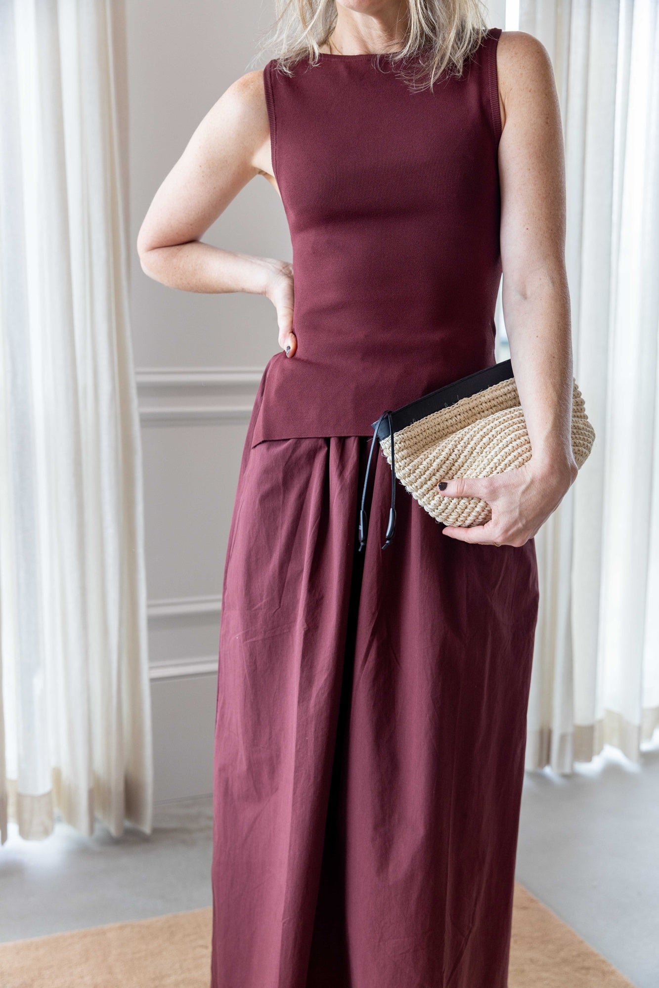 Close up of a Woman wearing the Onia boatneck dress in dark cherry holding the hunting season clutch and standing on a tan rug in front of white curtains.