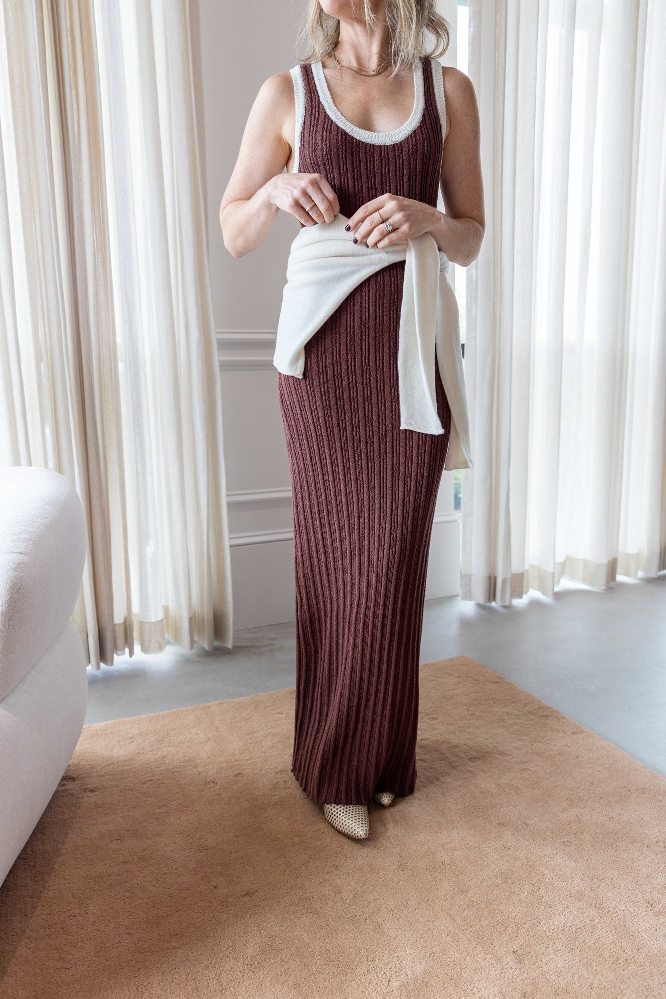 Close up of Woman wearing the Onia Cotton Boucle Scoop Tank Dress and sablyn lance sweater carrying a hunting season clutch standing on a tan carpet in front of white curtains.