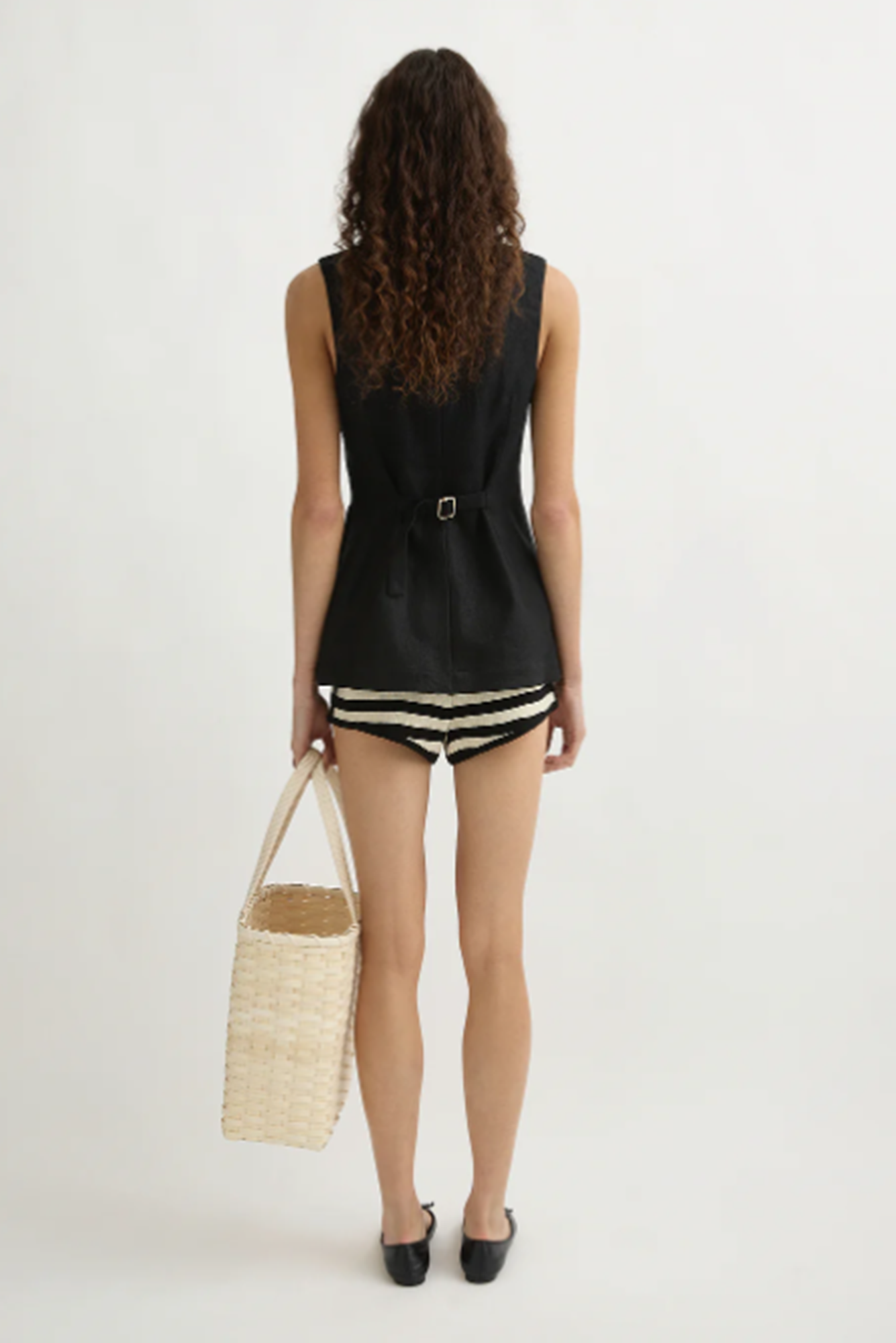 Woman wearing a black sleeveless top and striped shorts, holding a woven bag on a white background