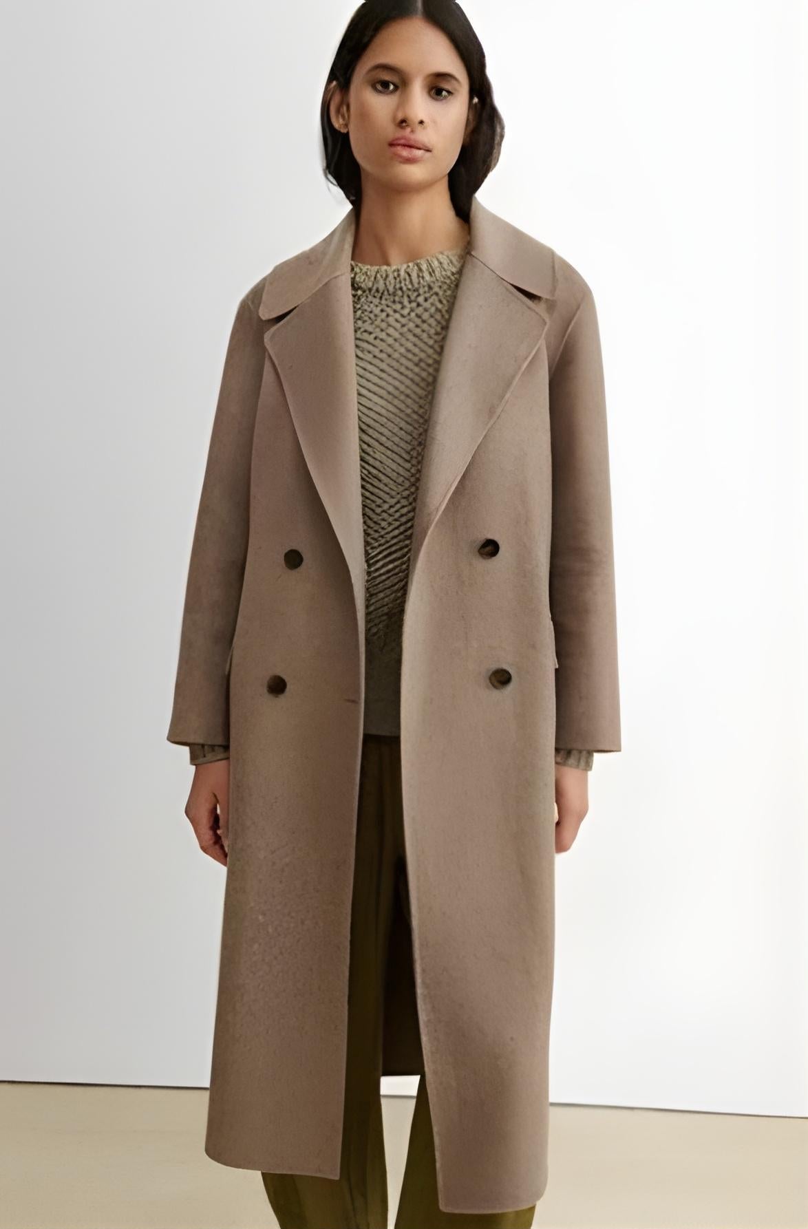 A woman modeling a long taupe-brown double-breasted overcoat over a textured knit sweater and olive green trousers, set against a plain light gray studio background.