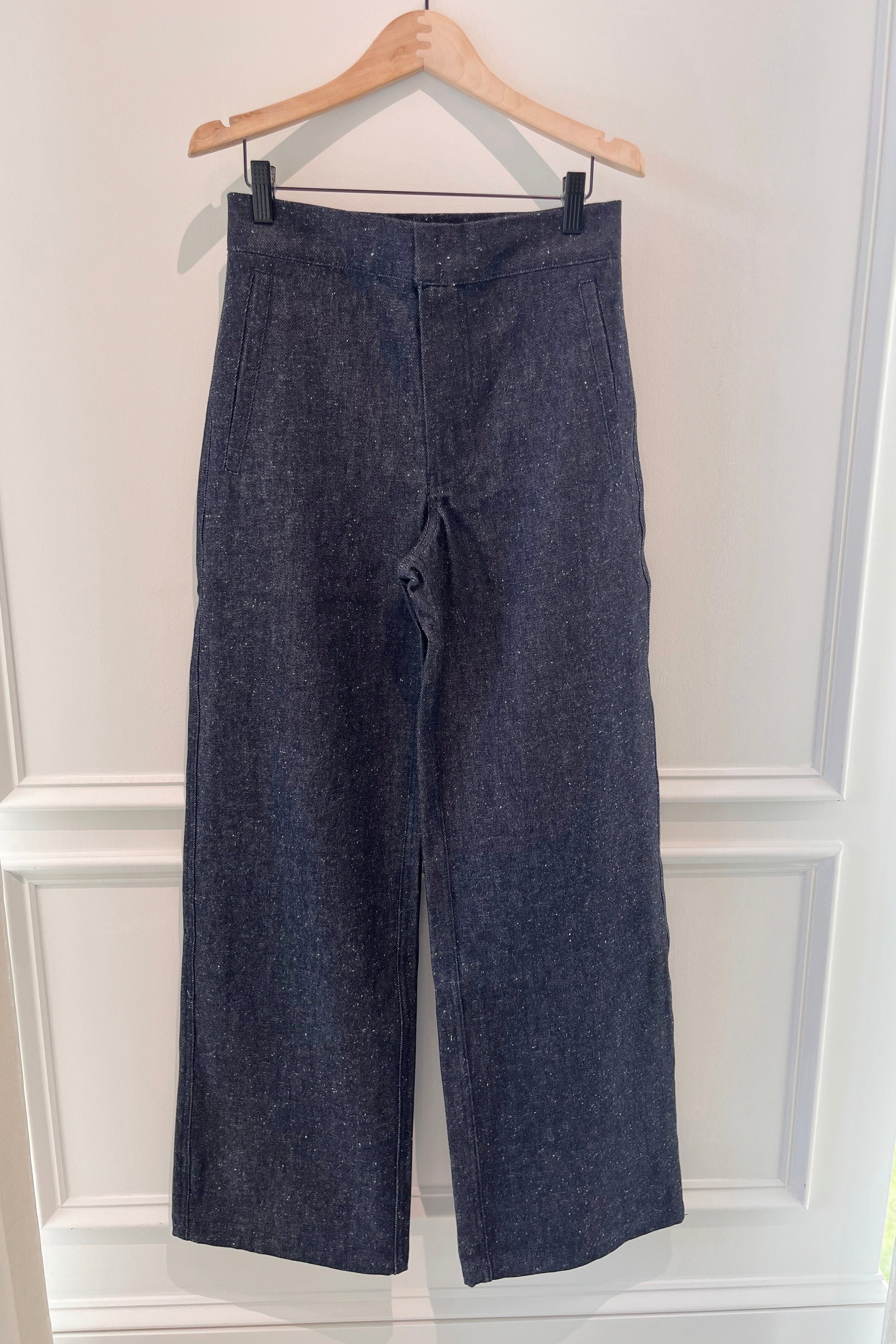 A pair of dark blue wide-leg cropped pants hanging from a wooden hanger against a white paneled wall.