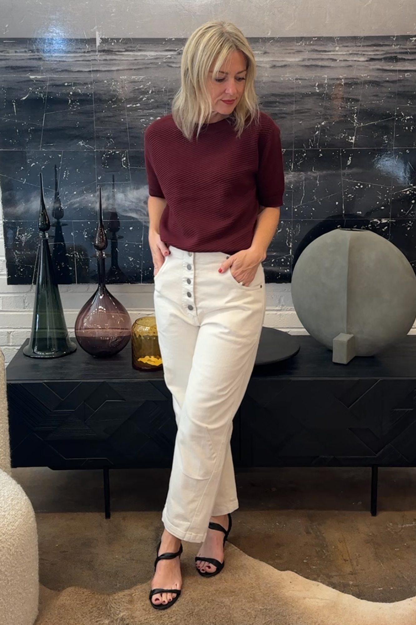 Woman in burgundy top and white pants standing in a room with decorative vases and sculptures.