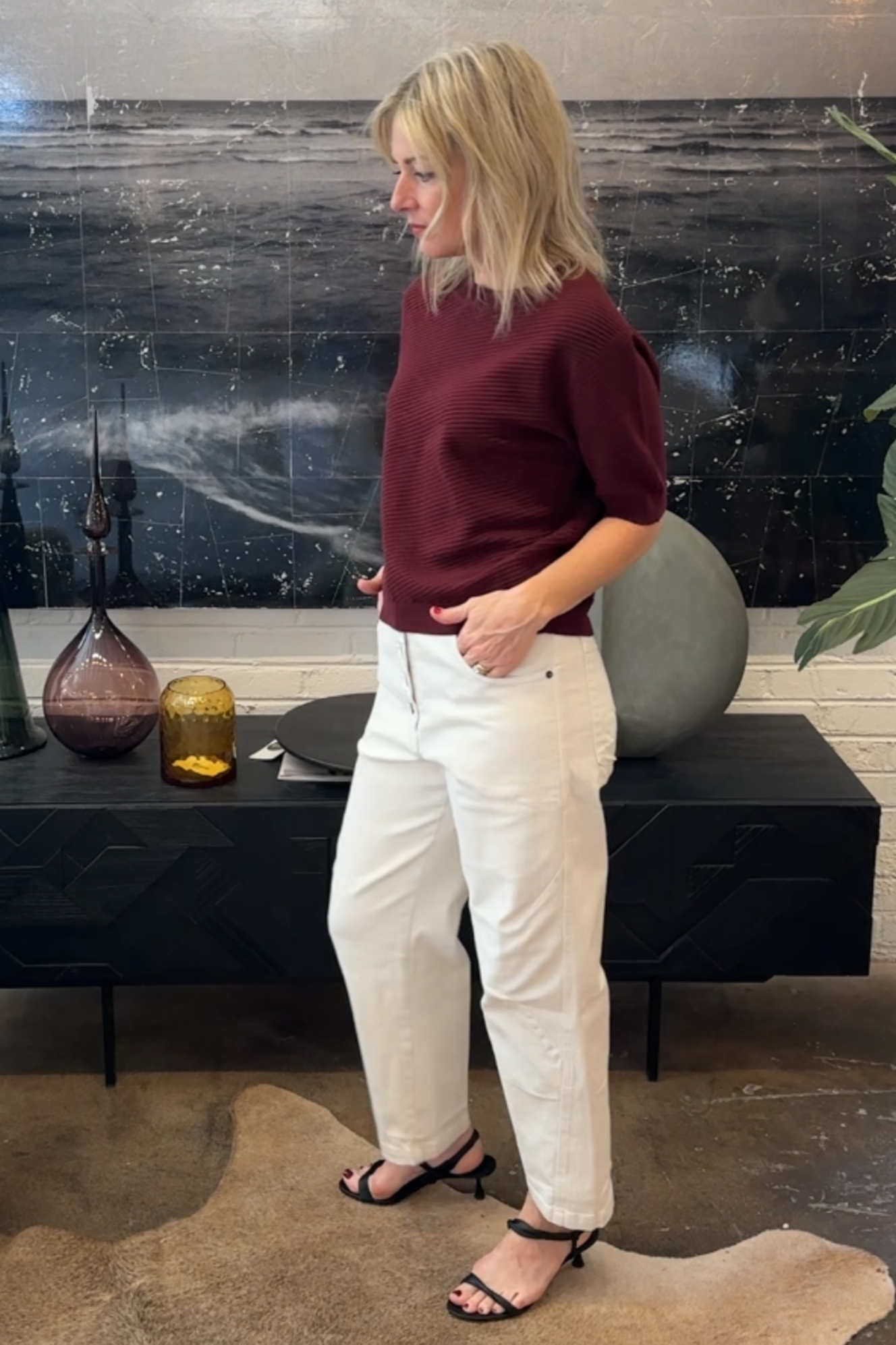 Woman in a burgundy sweater and white pants standing in a room with decorative elements.