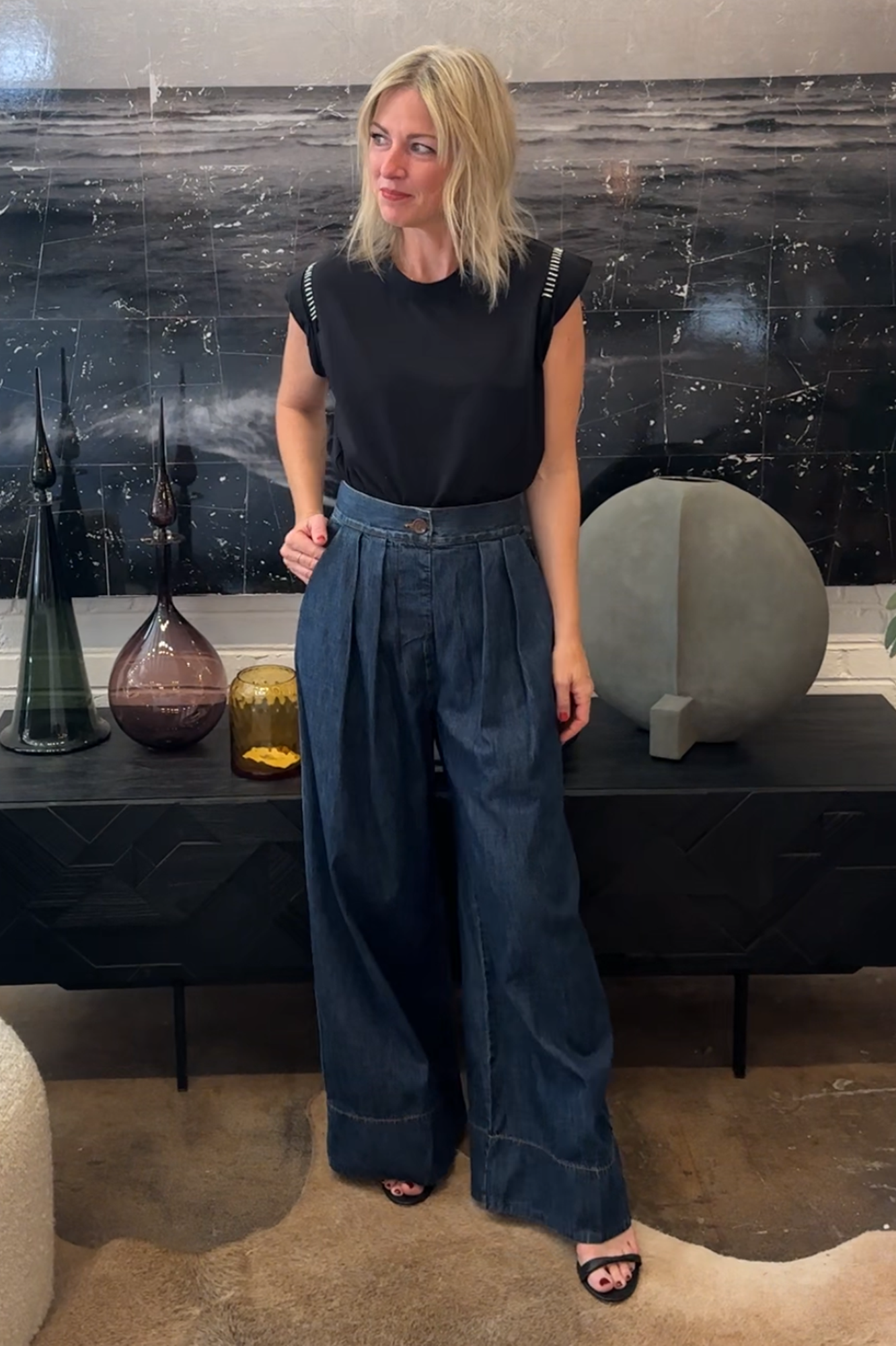 Woman wearing a black top and blue jeans standing in a room with decorative vases and a painting.
