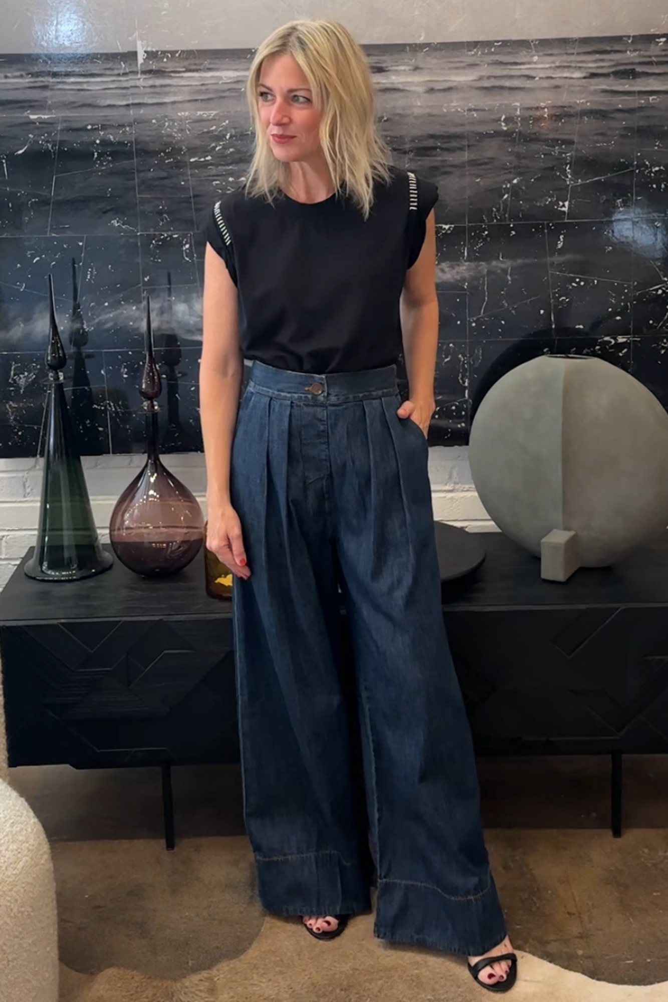 Woman in black top and blue pants standing in a room with decorative items on a shelf.