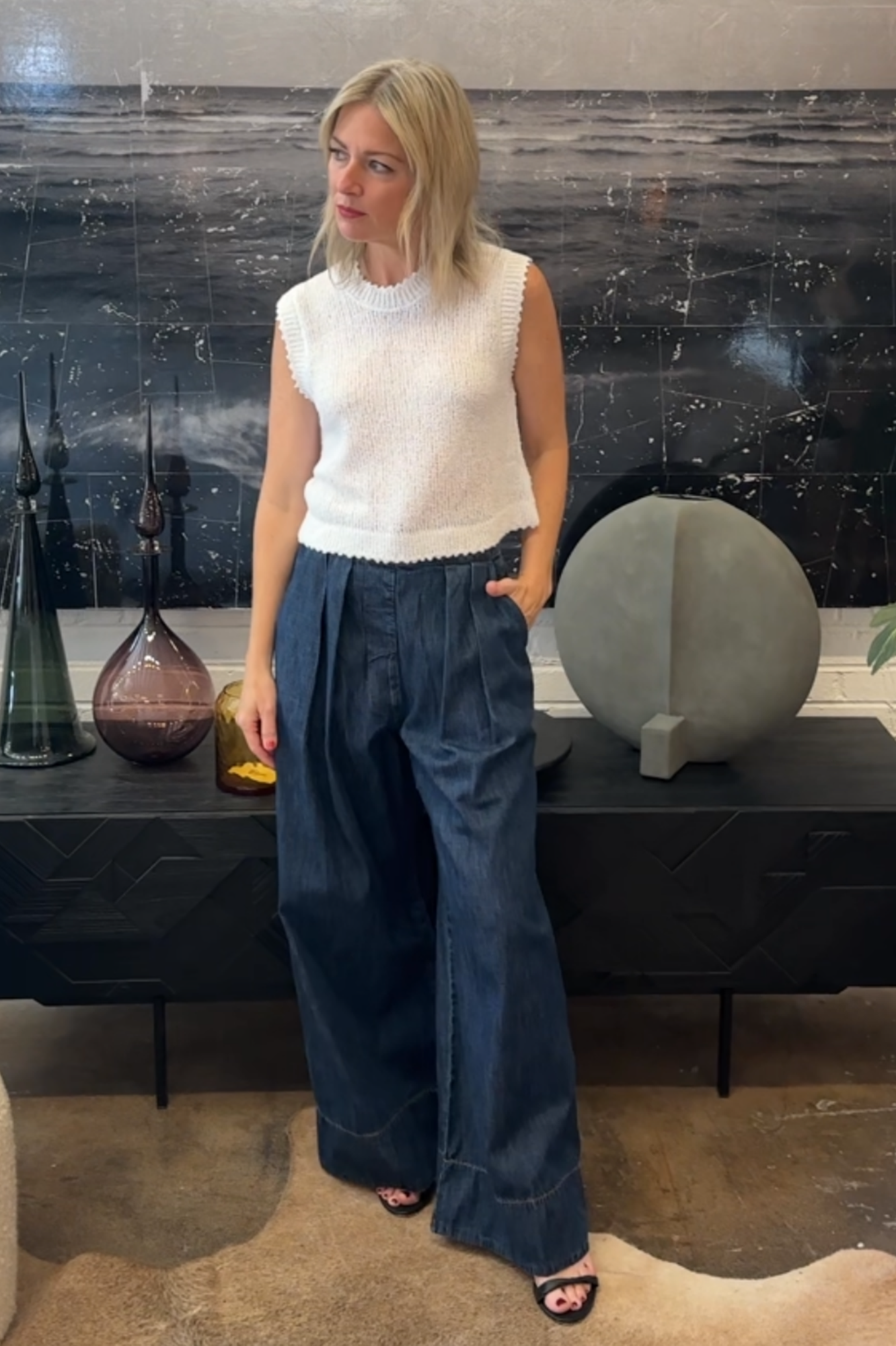 Woman in a white sleeveless top and blue wide-leg pants standing in front of decorative vases.