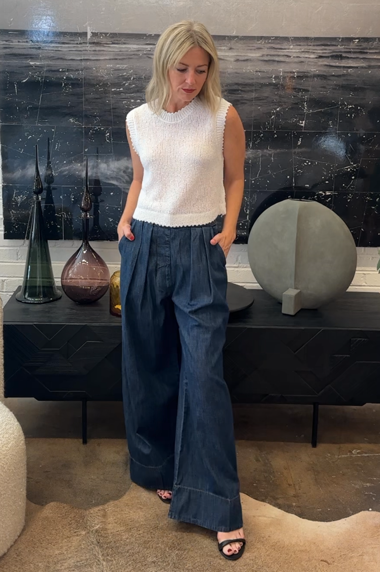 Woman in a white sleeveless top and blue jeans standing in a room with decorative items on a shelf.
