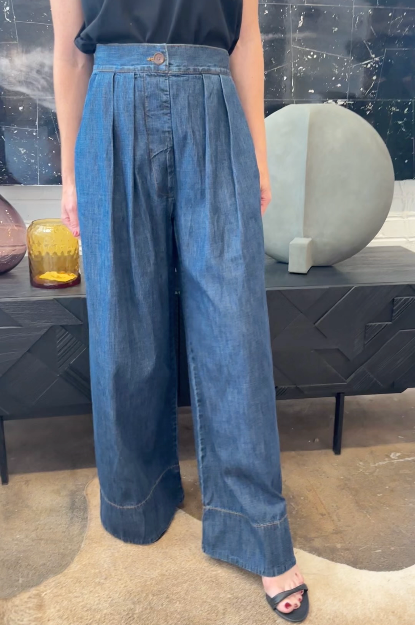 Person wearing blue wide-leg jeans standing in a room with decorative items on a shelf.