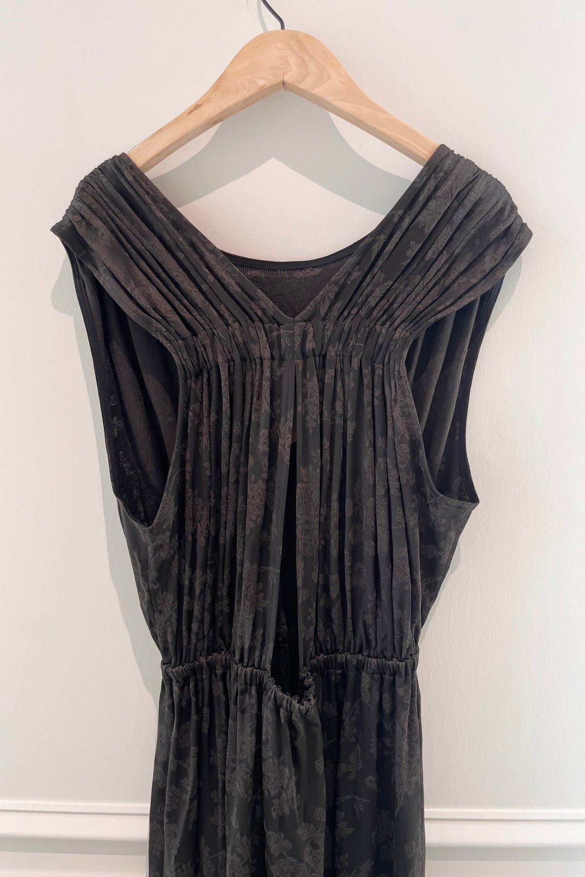 A dark brown textured velvet sleeveless dress with pleated detailing, hanging on a wooden hanger against a white wall.