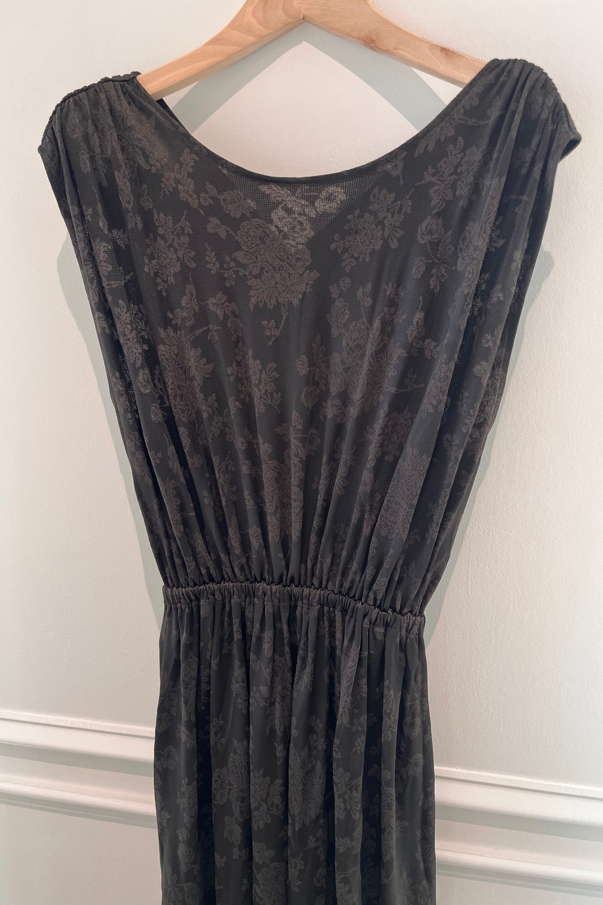A dark gray sleeveless dress with a subtle floral jacquard pattern, hanging on a wooden hanger against a white wall.