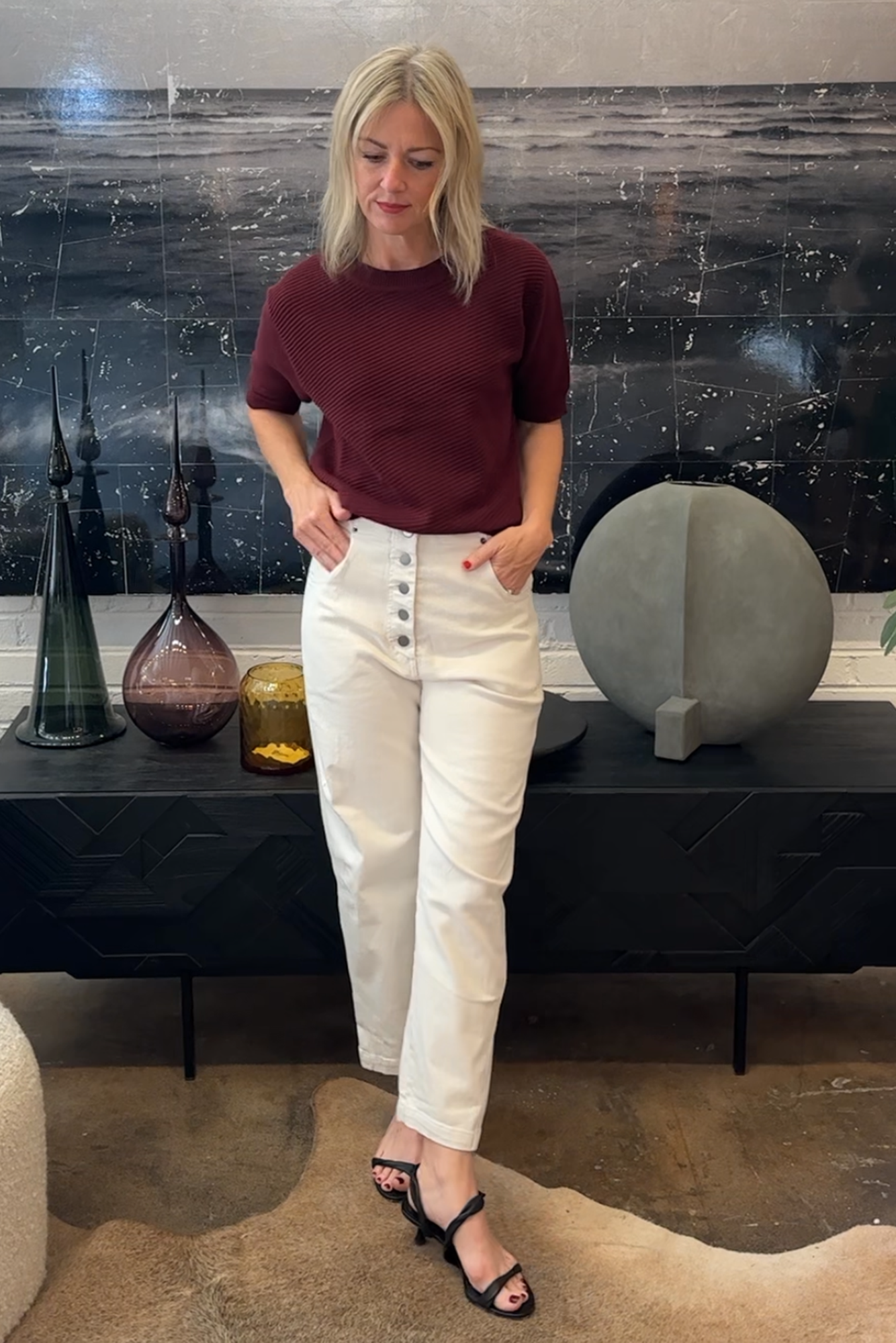 Woman in burgundy top and white pants standing in a room with decorative items on a shelf.