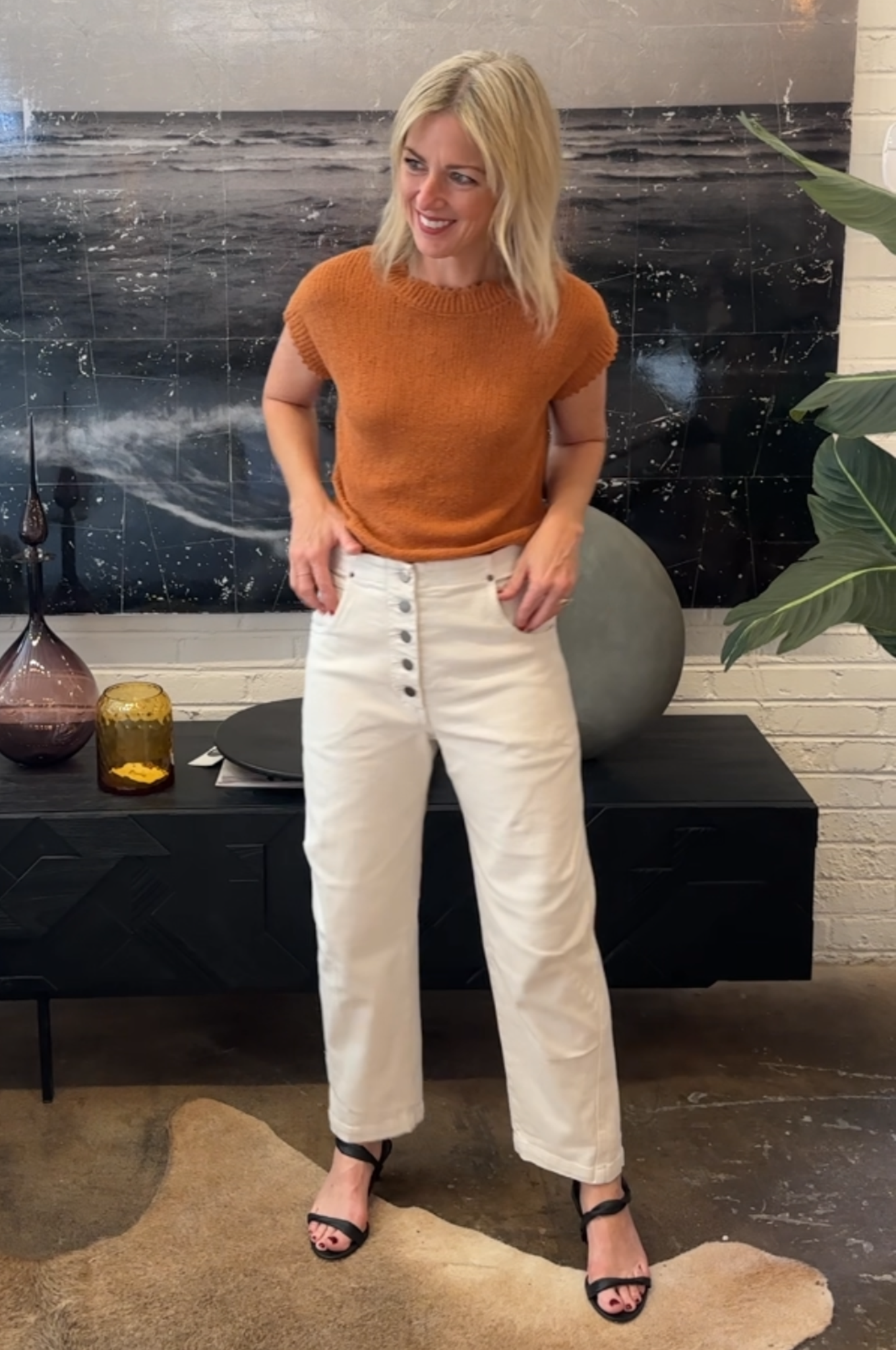 Woman in a rust-colored top and white pants standing in a modern interior setting.