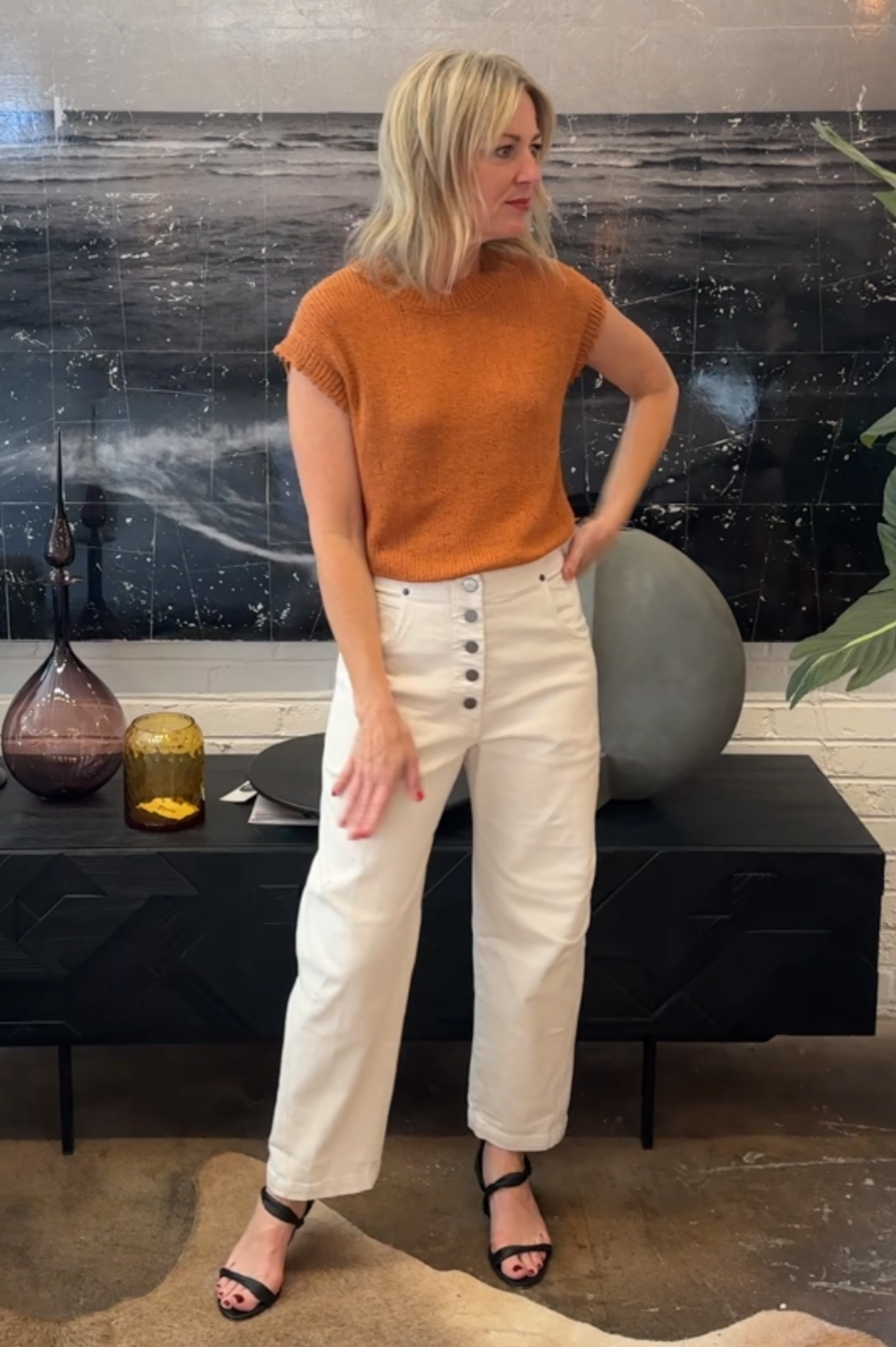Woman wearing an orange top and white pants standing in a room with a black wall and decorative items.