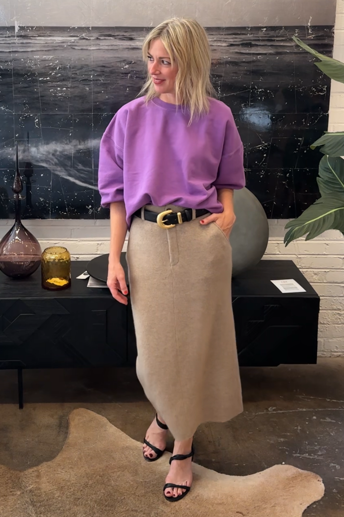 Woman in a purple sweater and beige skirt standing in a room with a black marble wall.