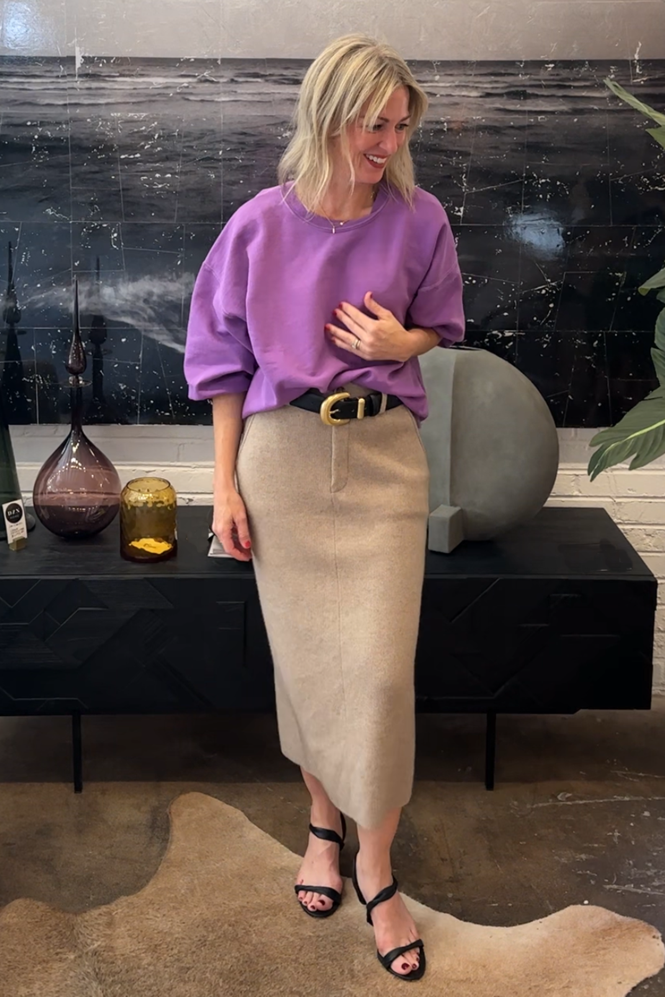 Woman in a purple sweater and beige skirt standing in a room with decorative items on a shelf.