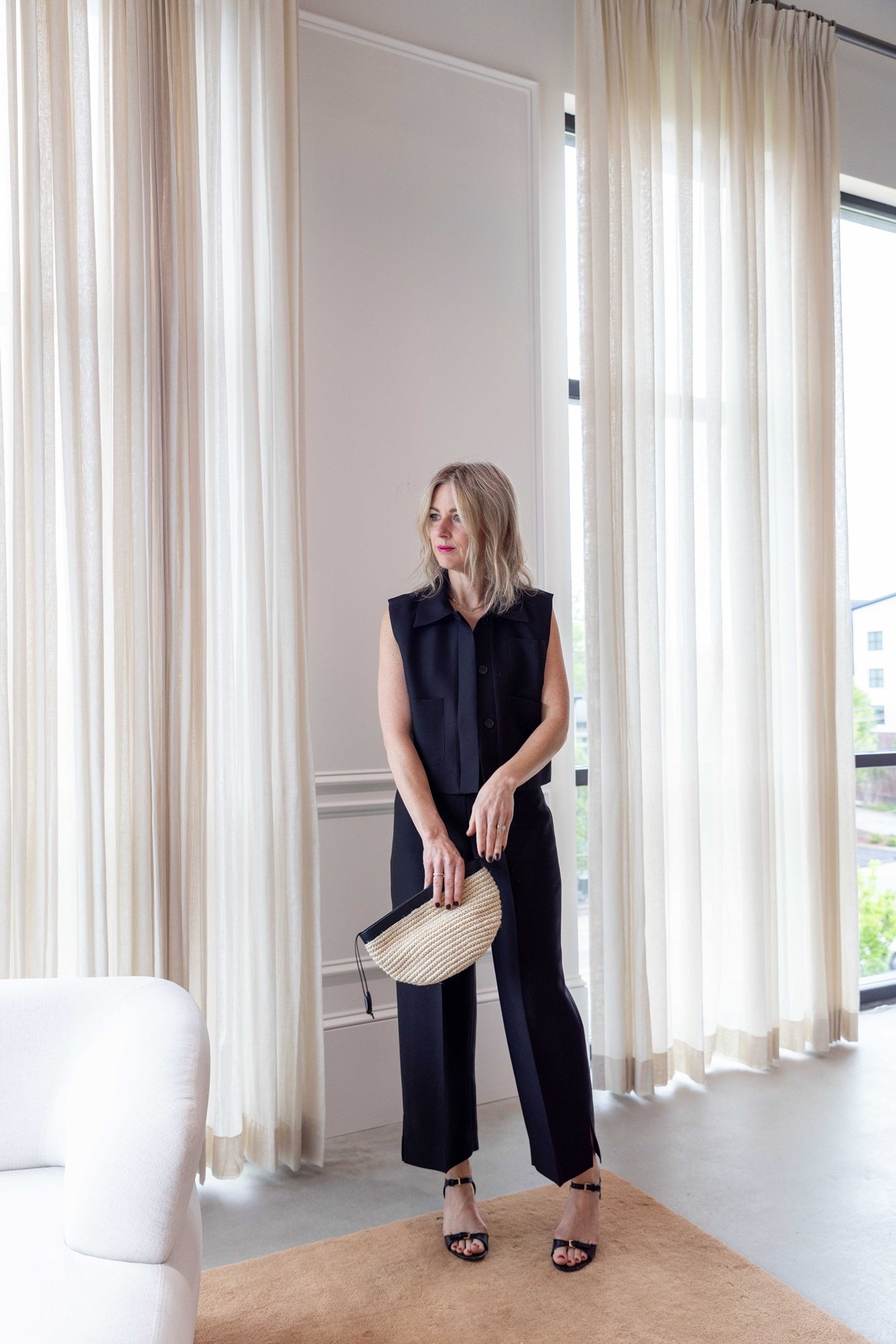 Woman wearing the rachel comey hanley vest, lam pant, and Sablyn Gloria V Neck Bias Cut Cami carrying hunting season clutch and standing in front of white curtains.