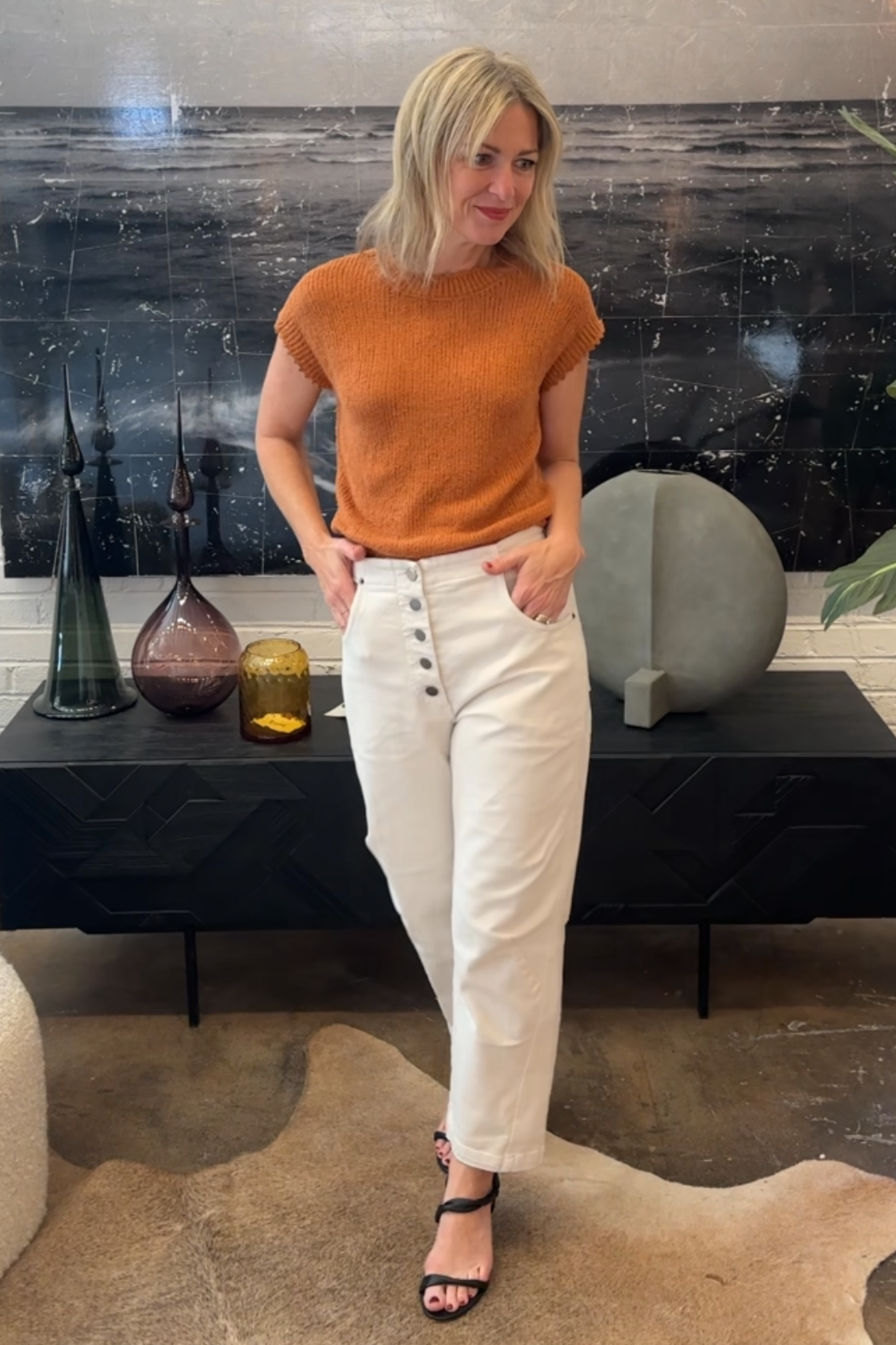 Woman wearing an orange top and white pants standing in a room with decorative items on a shelf.