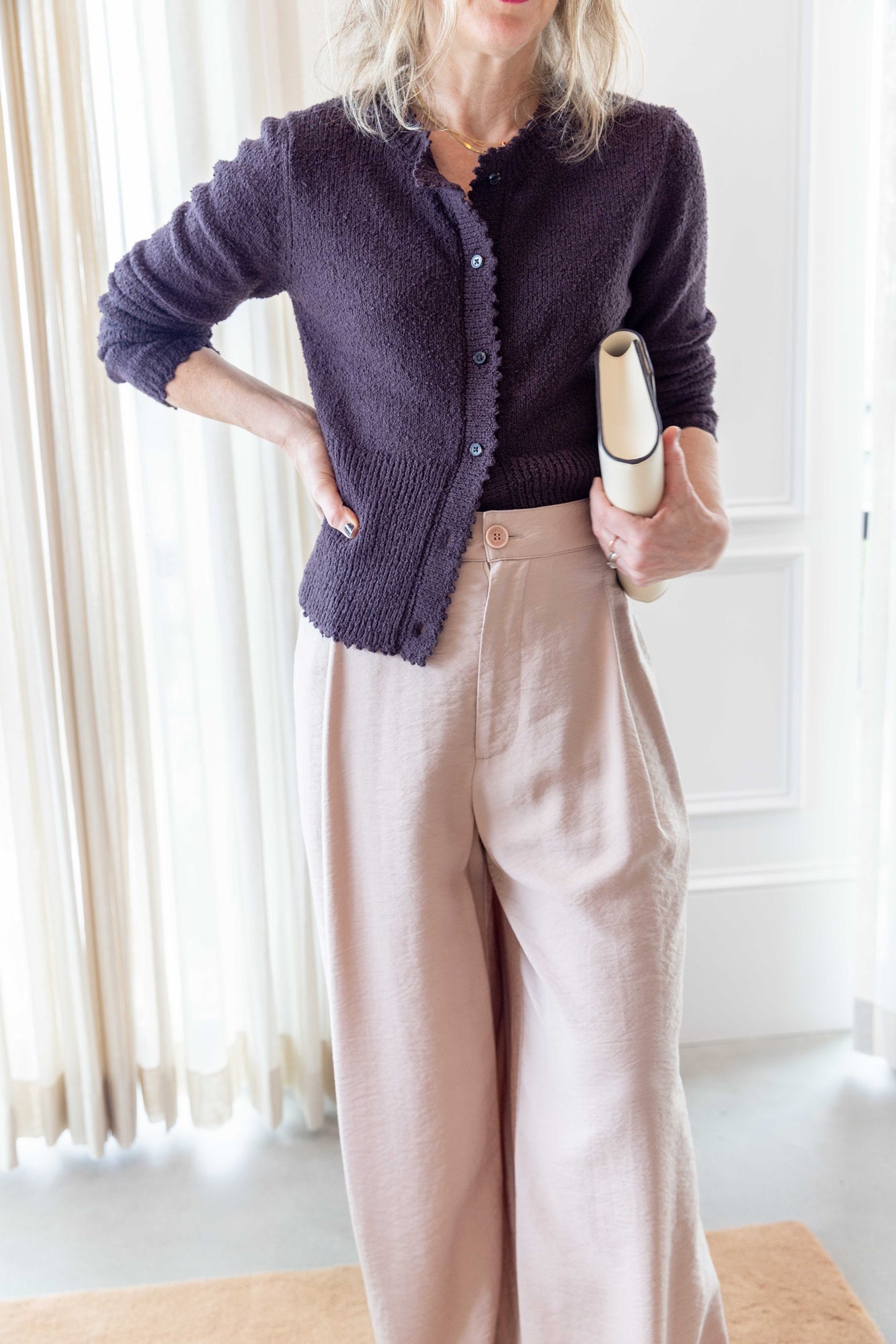 Close up of a Woman wearing Sablyn Sebastian B Relaxed Pleated Pant In color dune and the rachel comey monti cardigan carrying a hunting season clutch standing on a tan rug and in front of white curtains