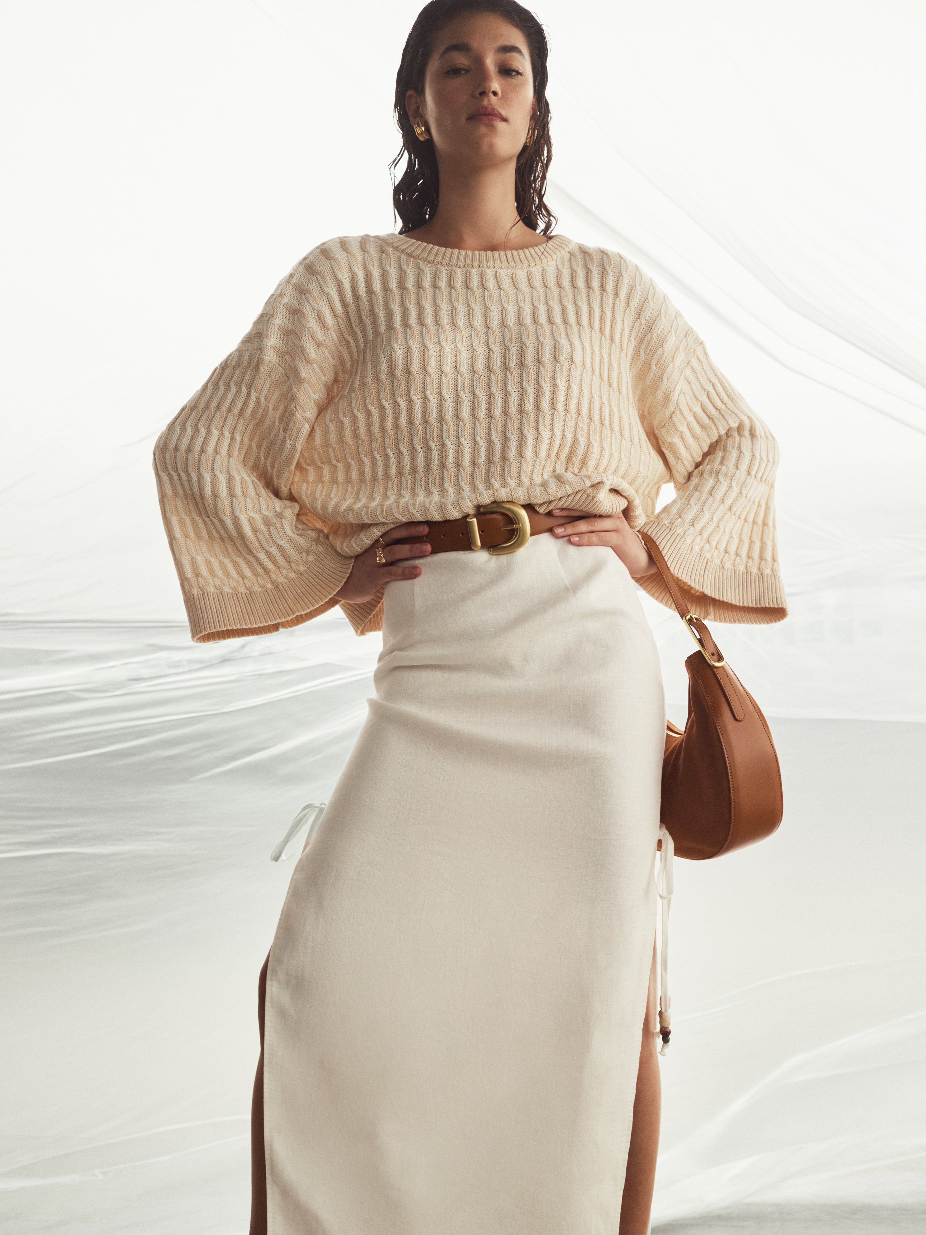 Woman wearing a beige sweater and white skirt with a brown handbag on a light background