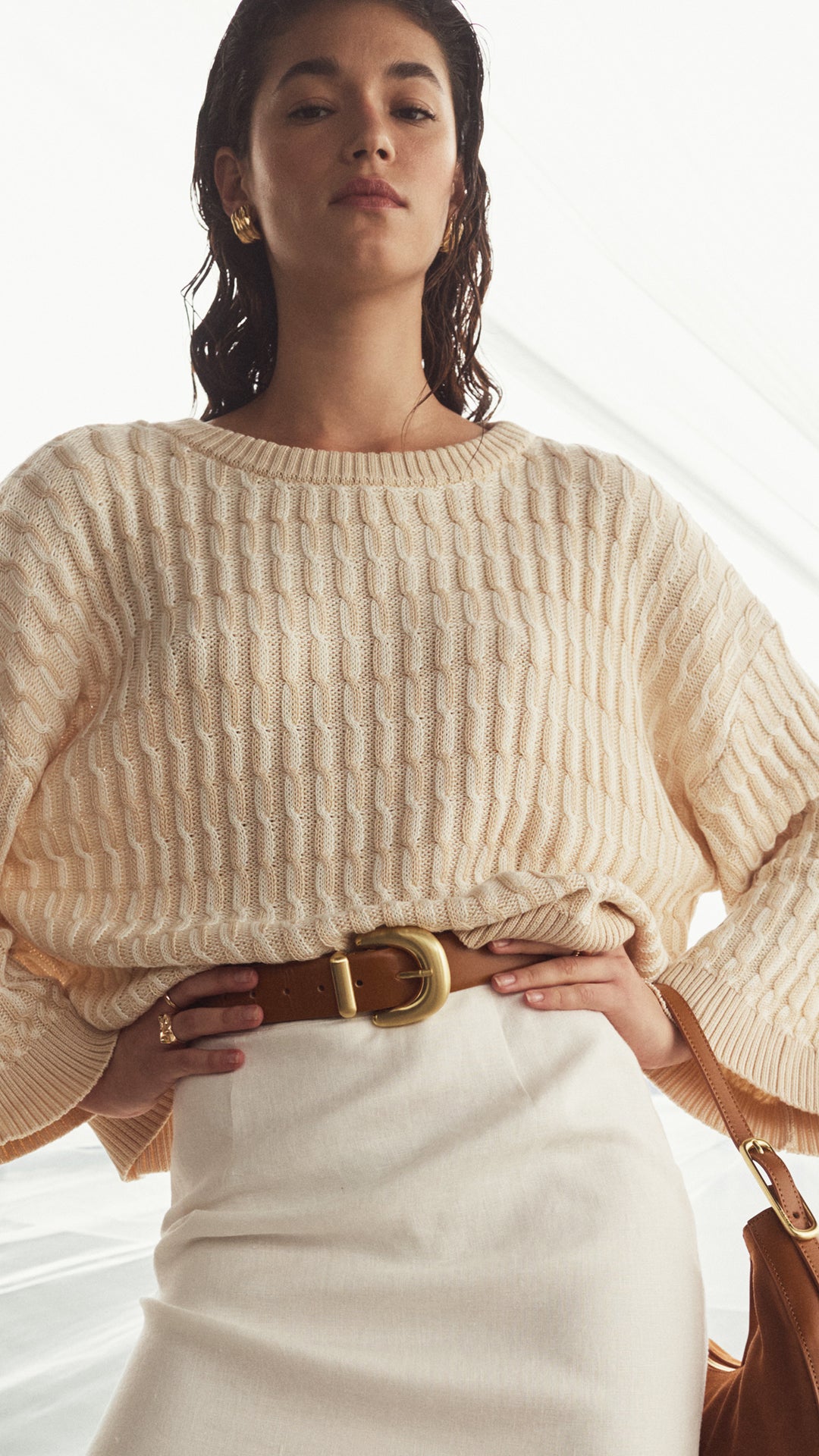 Woman wearing a beige ribbed sweater with a brown belt and white pants, standing against a neutral background.