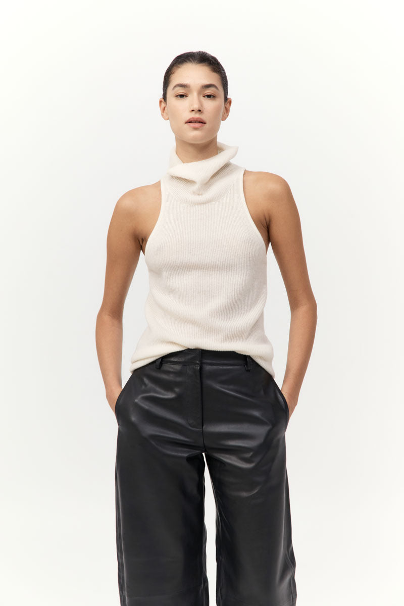 Woman wearing a cream sleeveless top and black pants on a white background