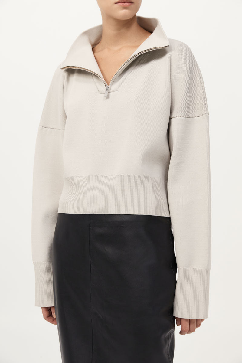Person wearing a light gray sweater with a high collar and black pants on a white background