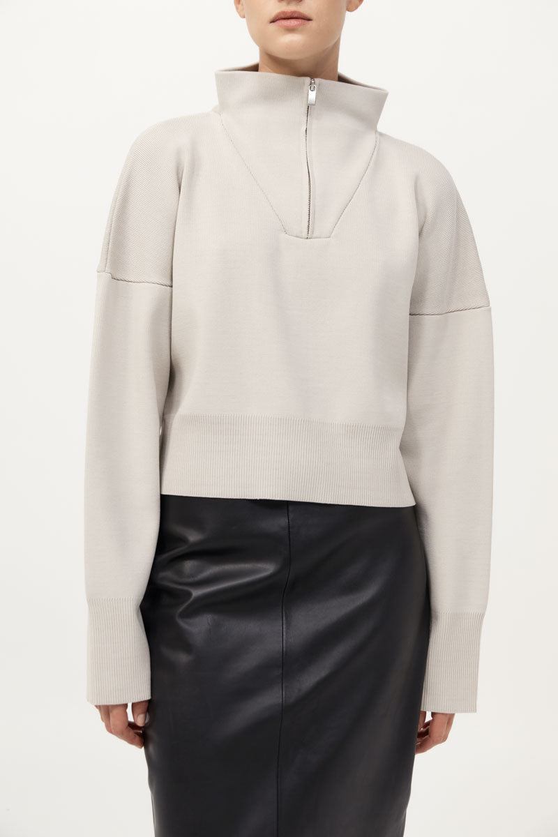 Person wearing a light gray sweater with a high collar and black leather pants on a white background