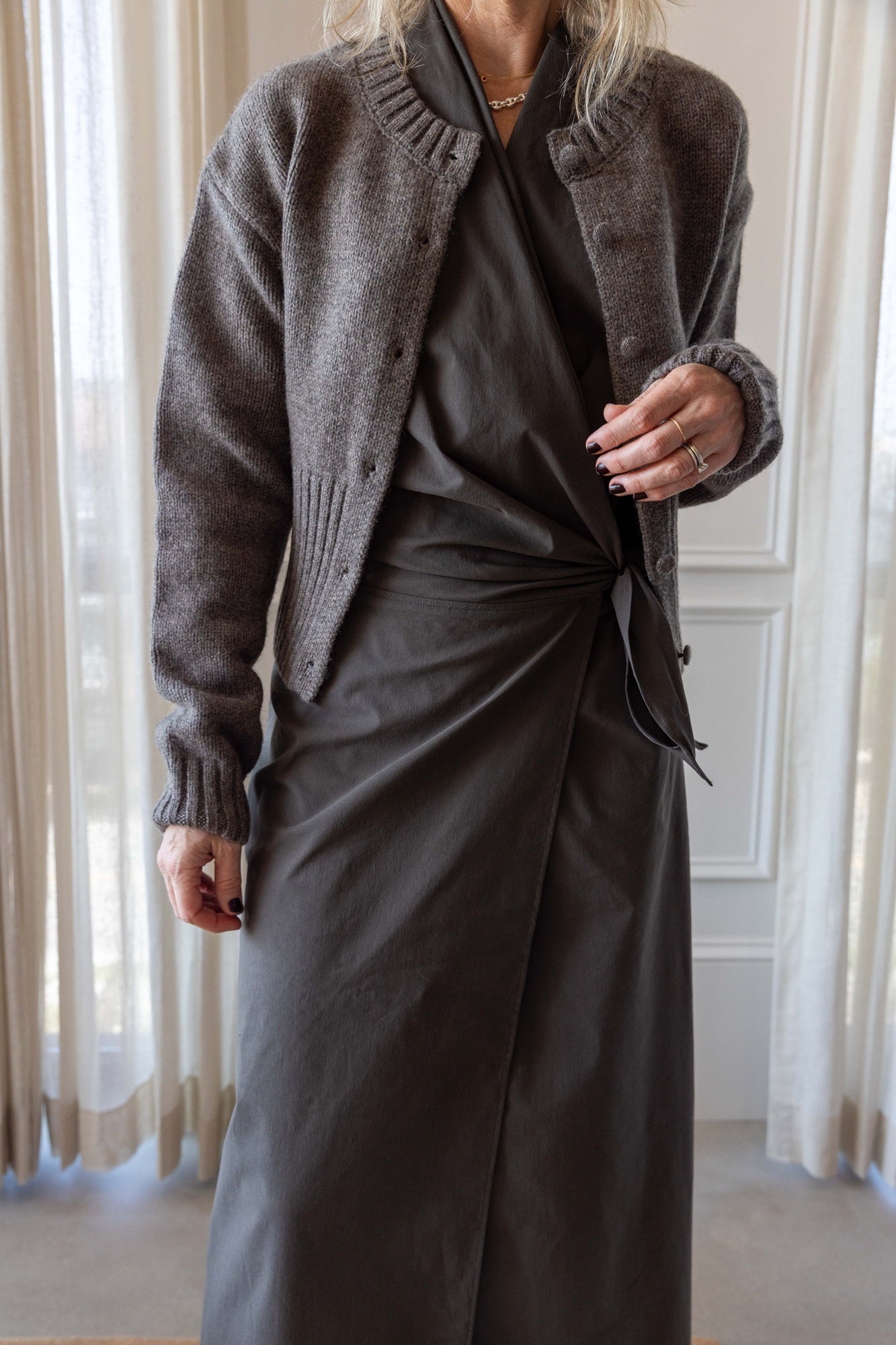 Woman wearing the St Agni Halter Wrap Backless Dress and wool cardigan standing against white curtains