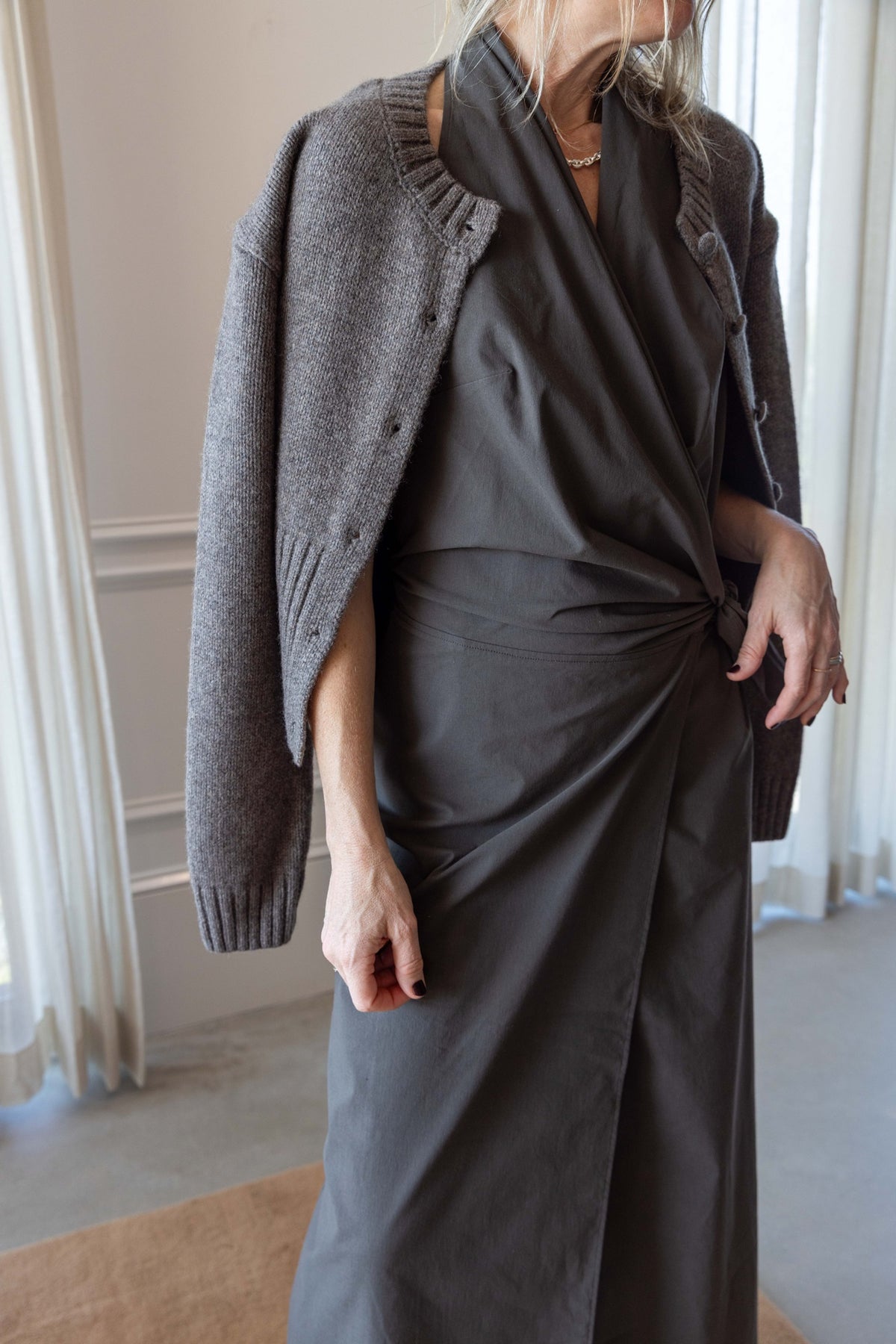 Close up of a woman wearing the St Agni Halter Wrap Backless Dress and wool cardigan standing against white curtains