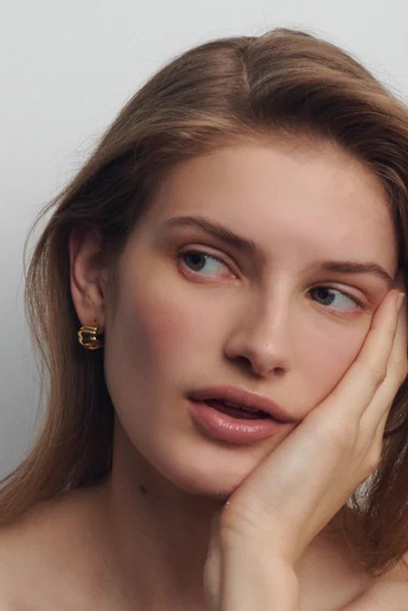 Woman with long brown hair and gold earrings against a plain background