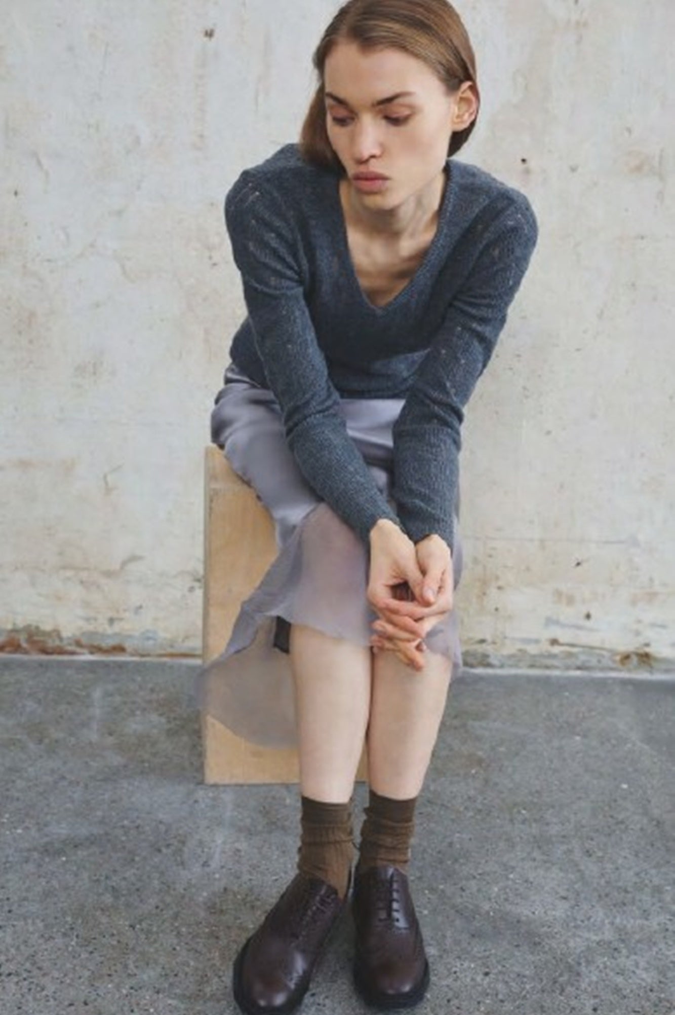 Woman sitting on a concrete block wearing a gray sweater, light skirt, brown socks, and dark shoes.