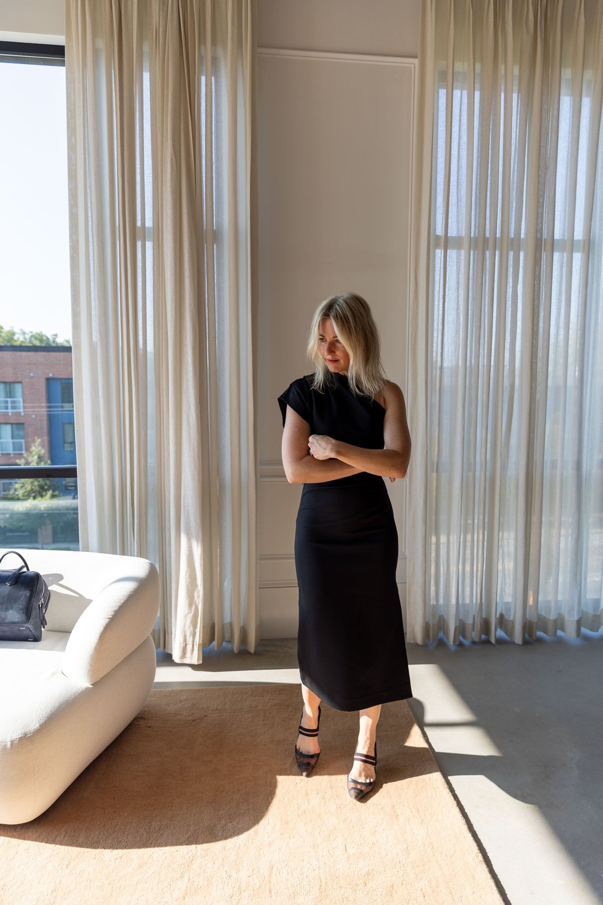 The Garment Glam Asymmetric Dress in black stretch cotton, worn full length in a sunlit room with black strappy heels