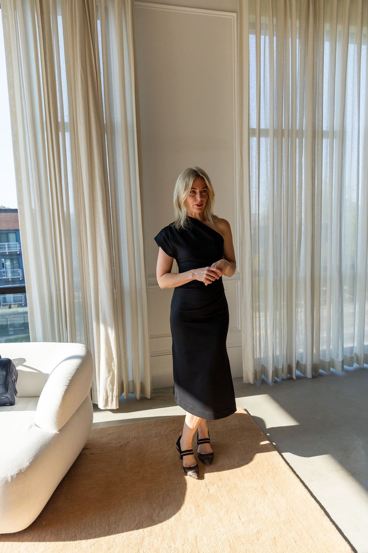 The Garment Glam Asymmetric Dress in black stretch cotton, worn front view showing the gathered shoulder and midi length