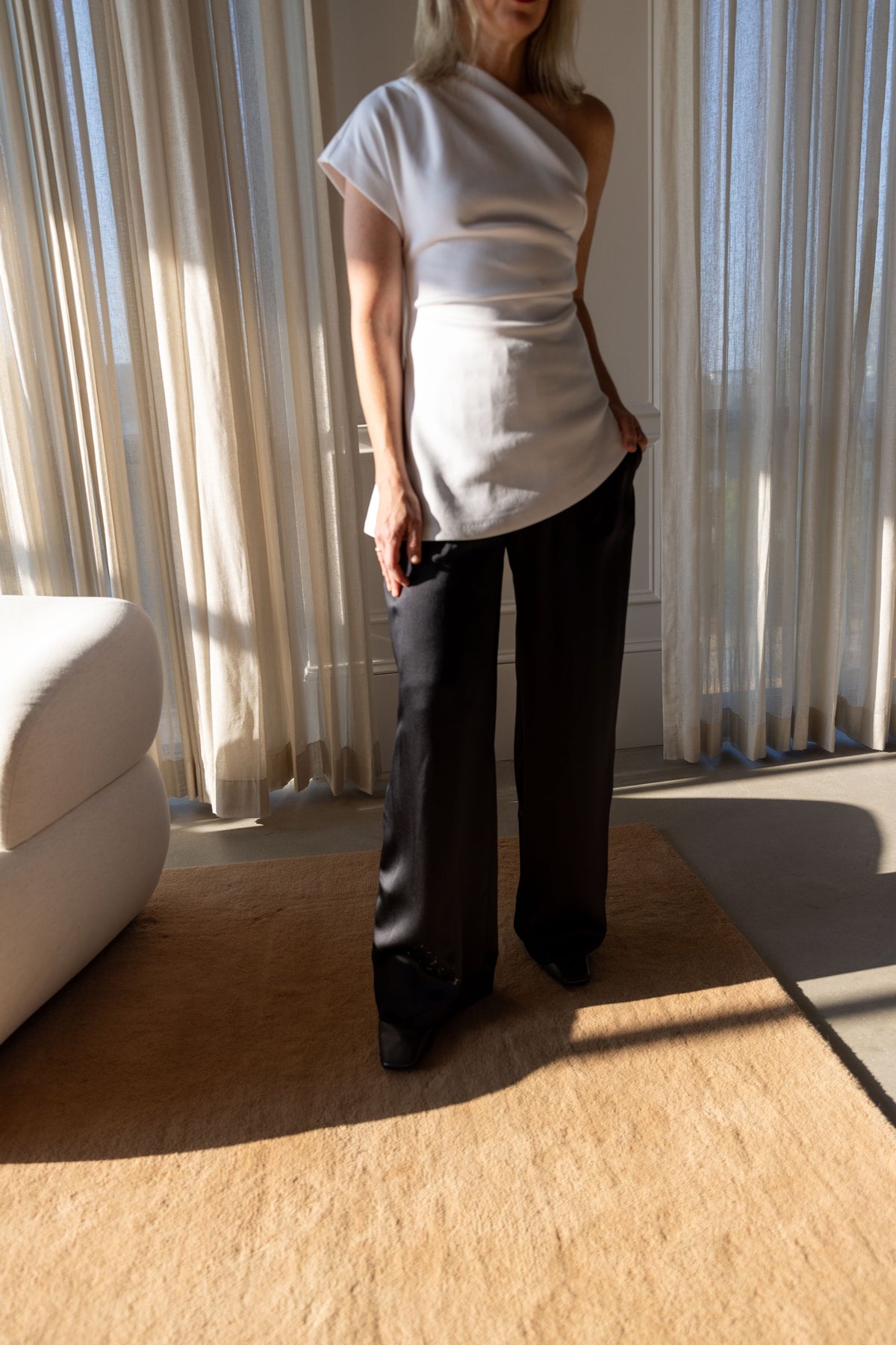 The Garment Glam Asymmetric Tee in cream stretch cotton, worn full length in a sunlit room with black wide-leg trousers