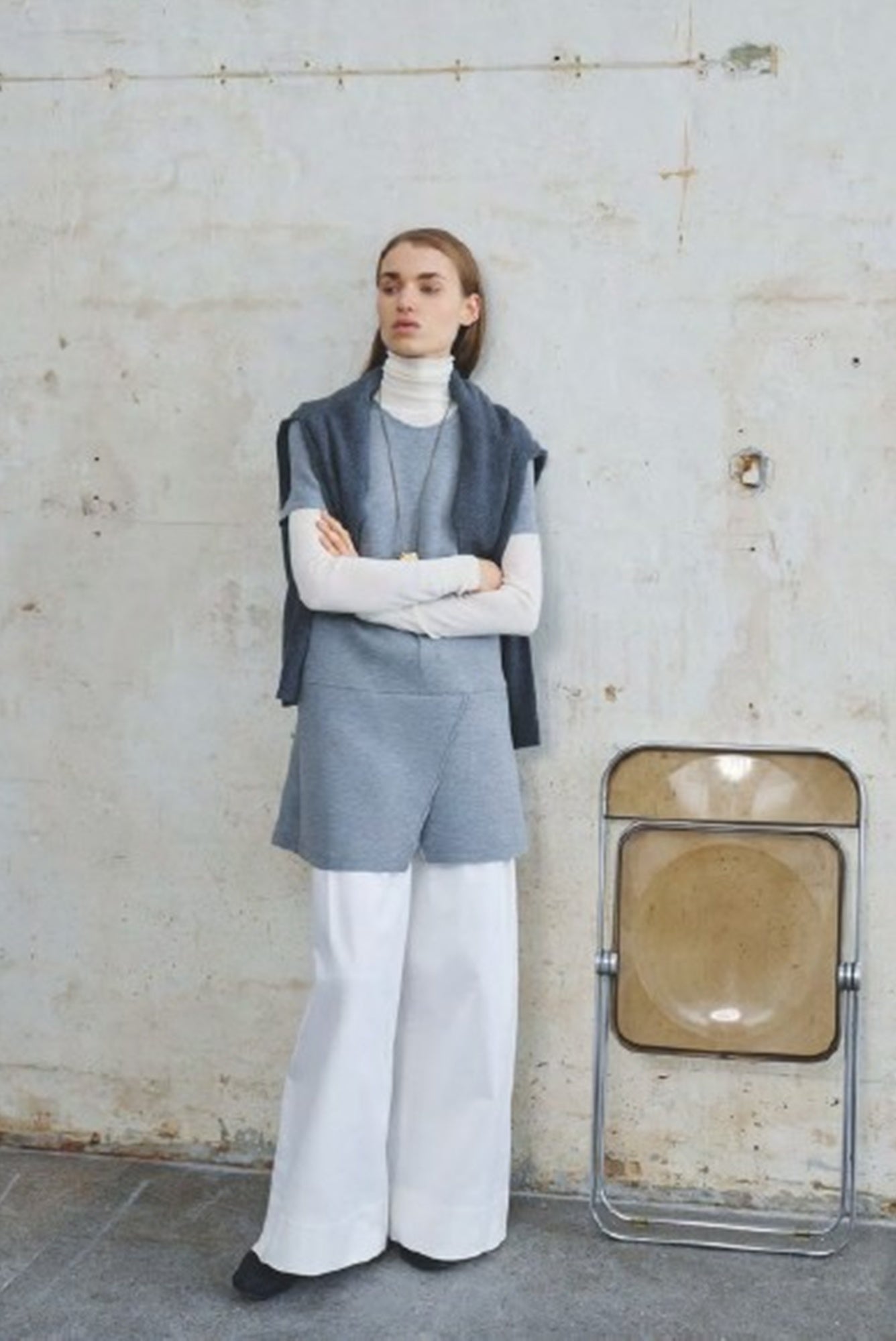 Person wearing a gray outfit with a white turtleneck and wide-leg pants, standing against a textured wall.