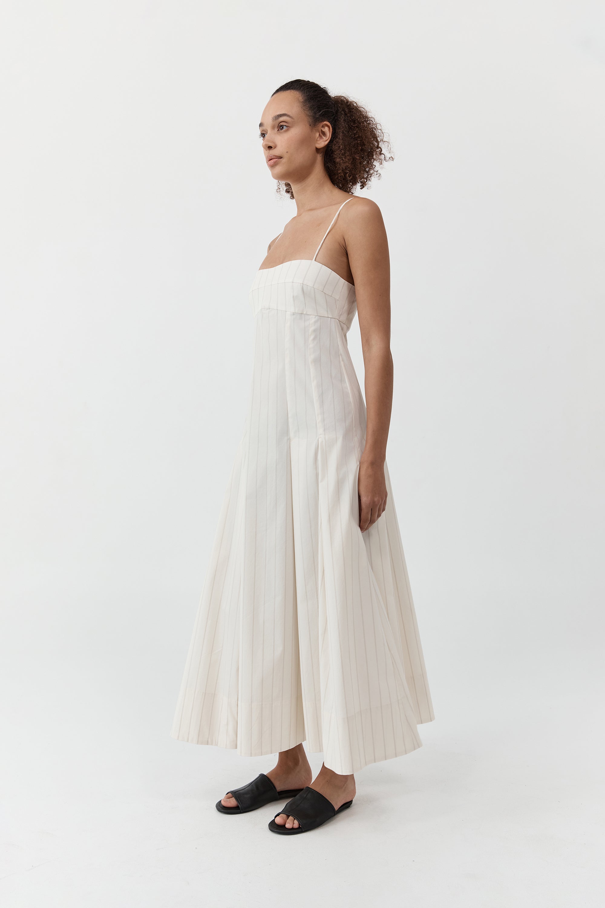Woman wearing the st. agni everyday cotton pleated dress in ecru stripe standing in front of a cream background