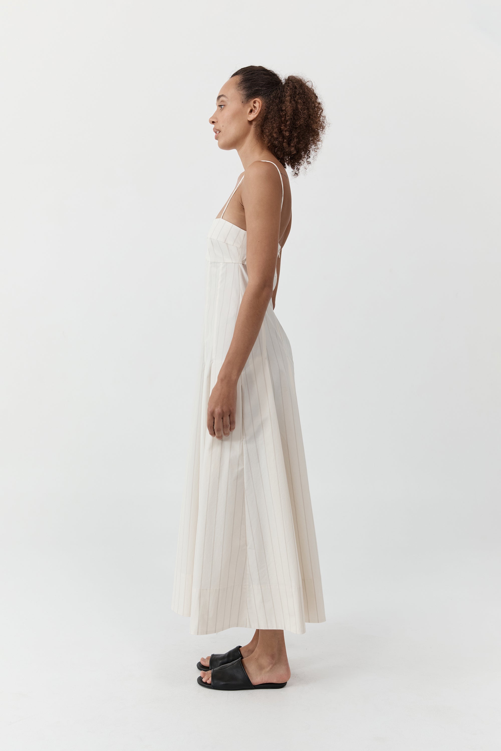 Side profile of a woman wearing the st. agni everyday cotton pleated dress in ecru stripe standing in front of a cream background