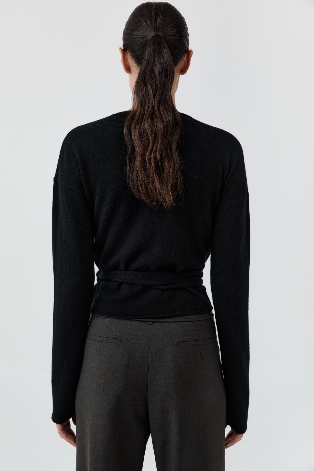 Backside of a woman wearing St Agni Baby Wool Wrap Top and gray pants standing against a white background