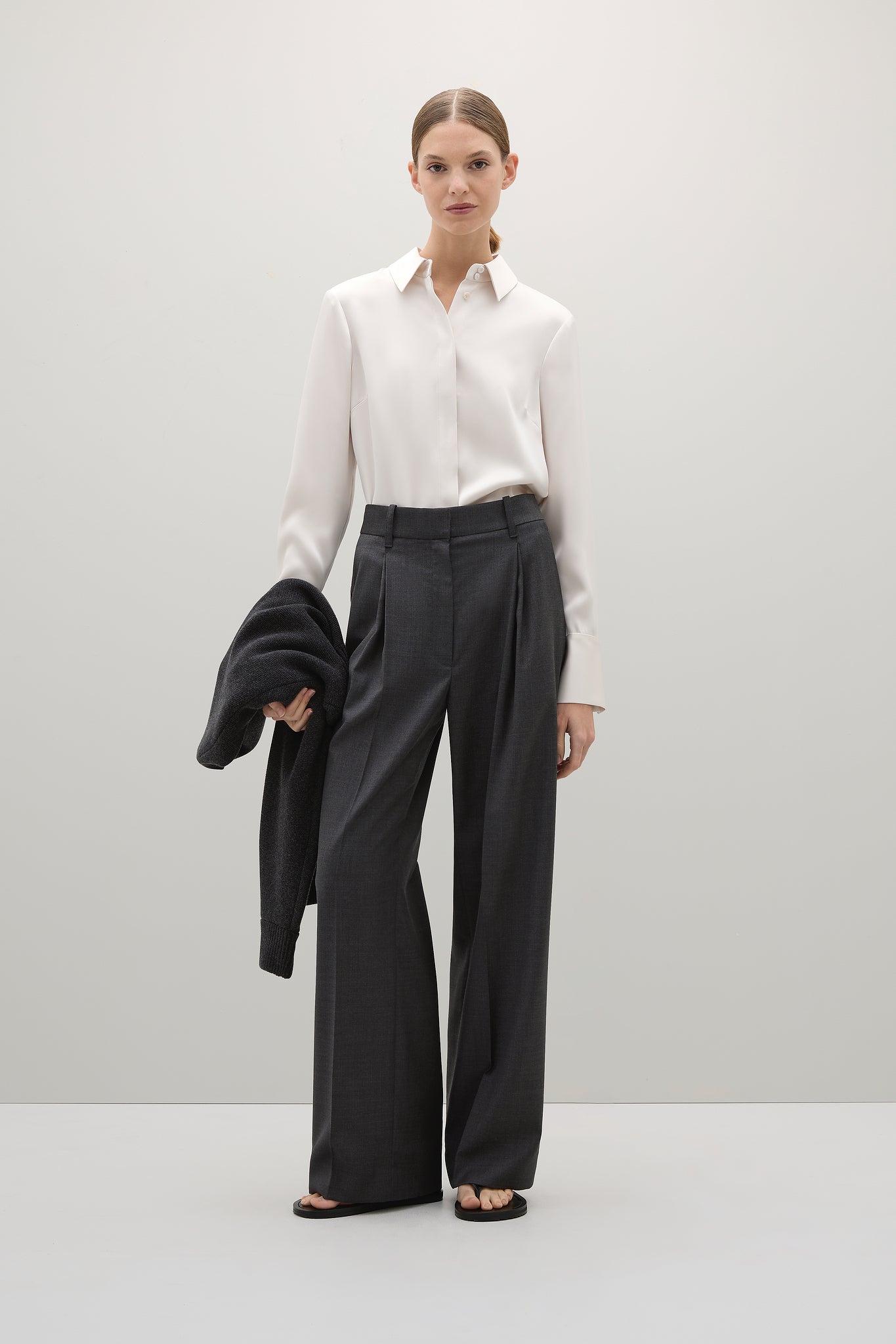 Woman wearing a white shirt and black pants with a black coat over her shoulder on a white background