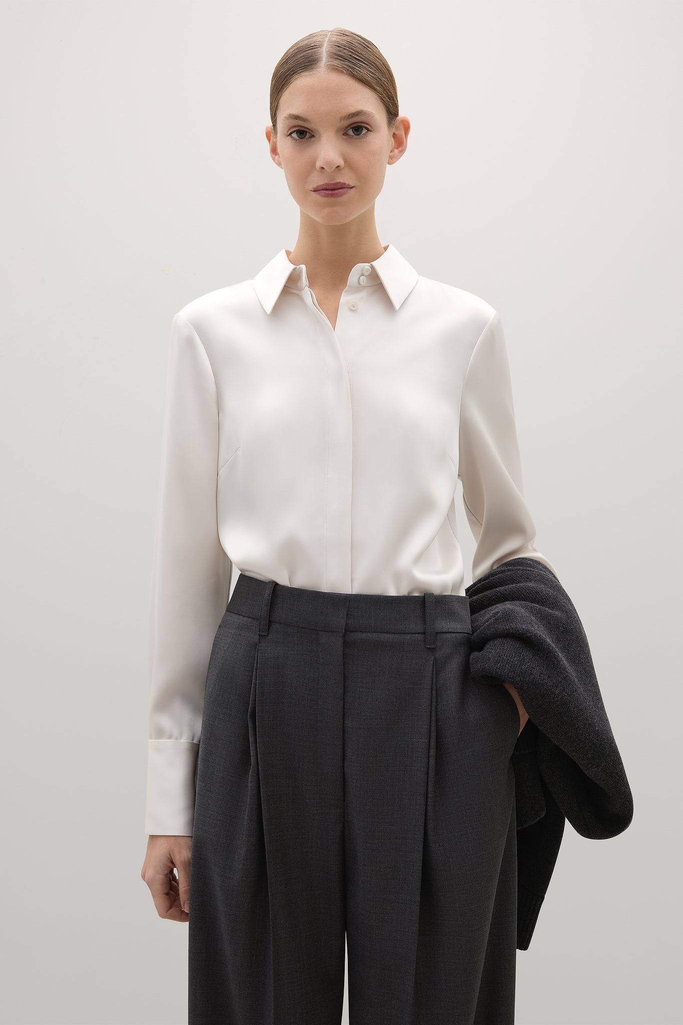 Woman wearing a white shirt and dark pants with a gray coat over her shoulder on a light background