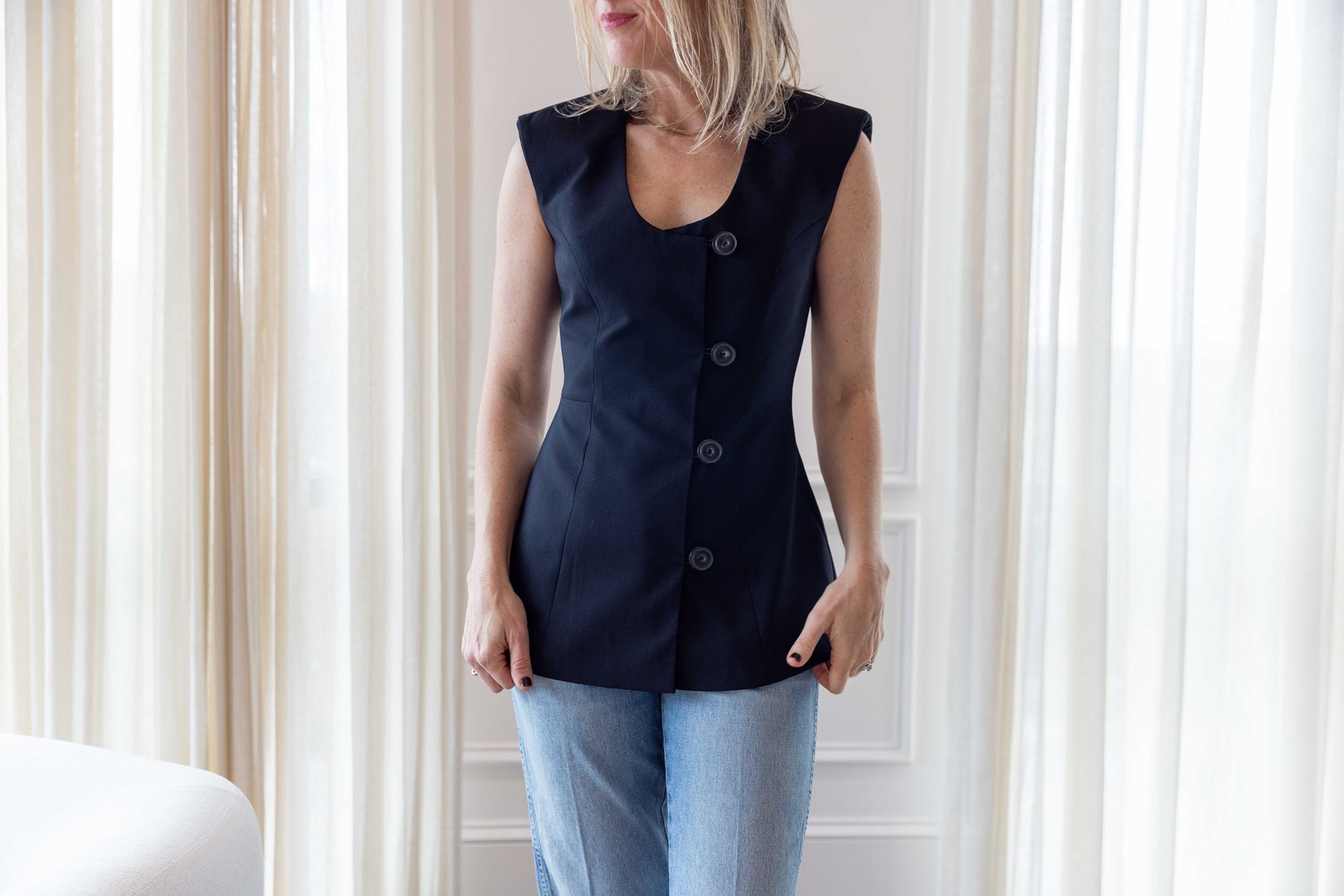 Close up of a woman wearing the house of dagmar sleeveless blazer and standing in a room with a white sofa and white curtains.