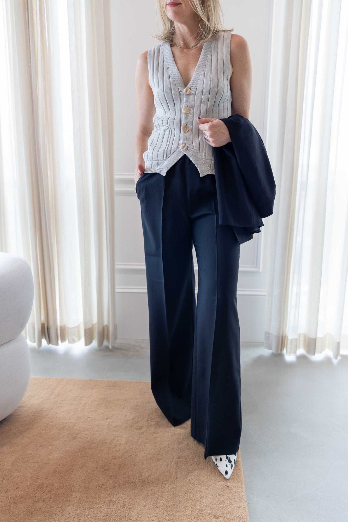 Close up image of a woman wearing the Amo marlowe vest and house of dagmar elastic wool trousers standing in a room with white curtains and a tan area rug.