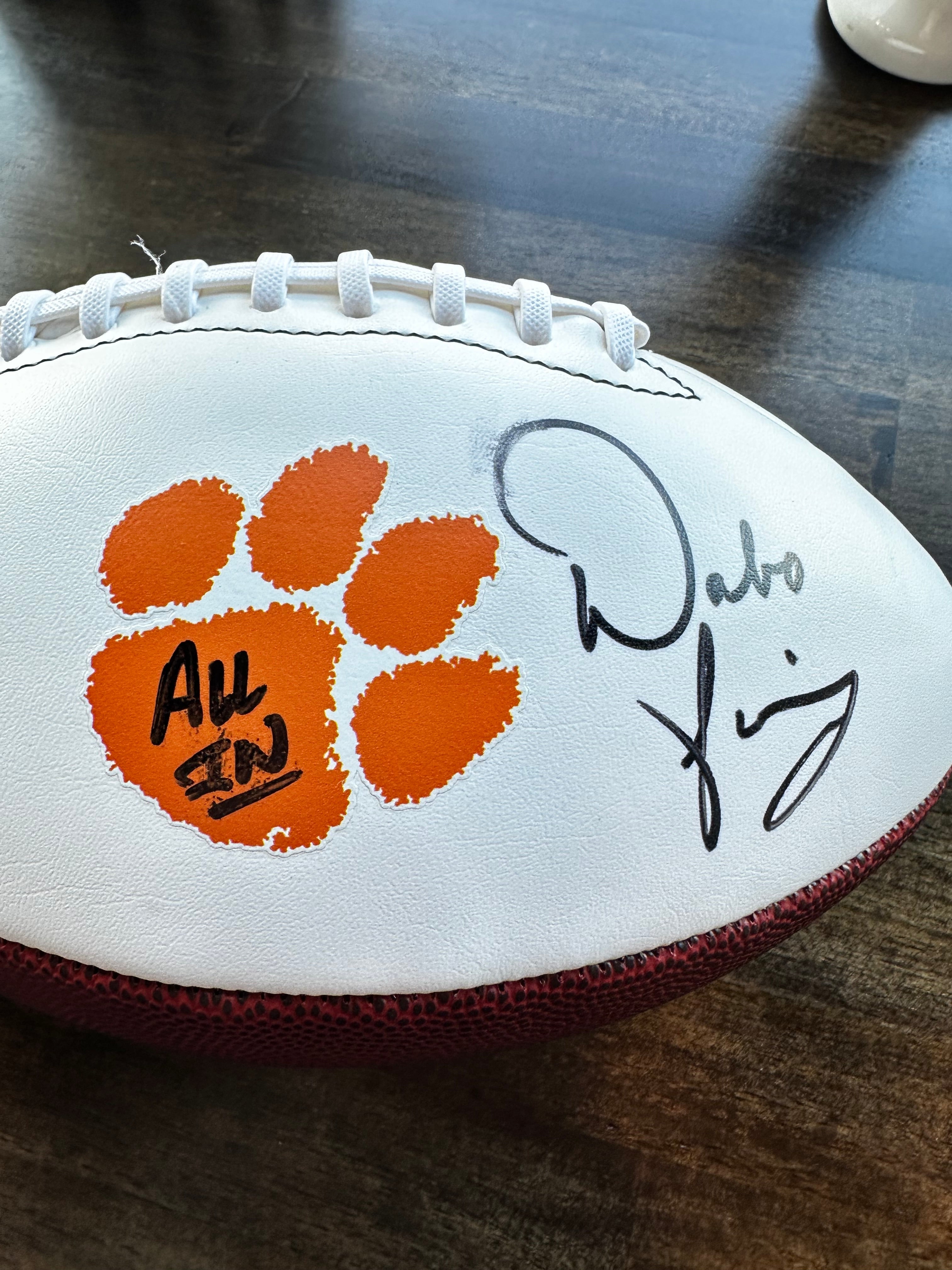 A white leather football with a maroon textured side, white laces, an orange paw print graphic, and black signatures on the front, photographed on a dark wooden surface.