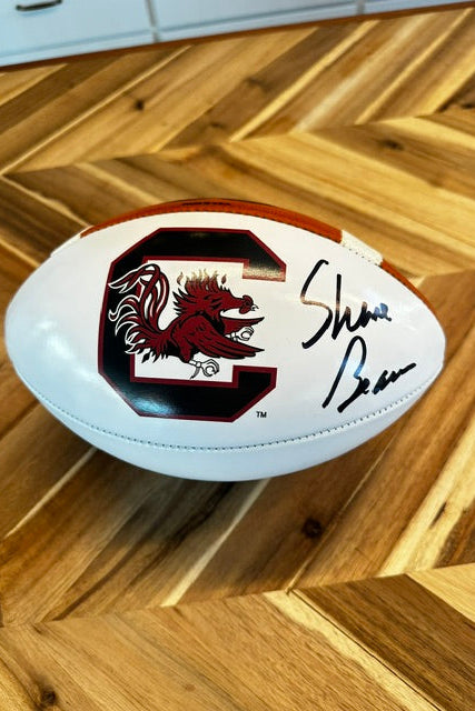 A white leather football with a red and black Southern Carolina logo and an autographed signature, resting on a wooden herringbone floor.